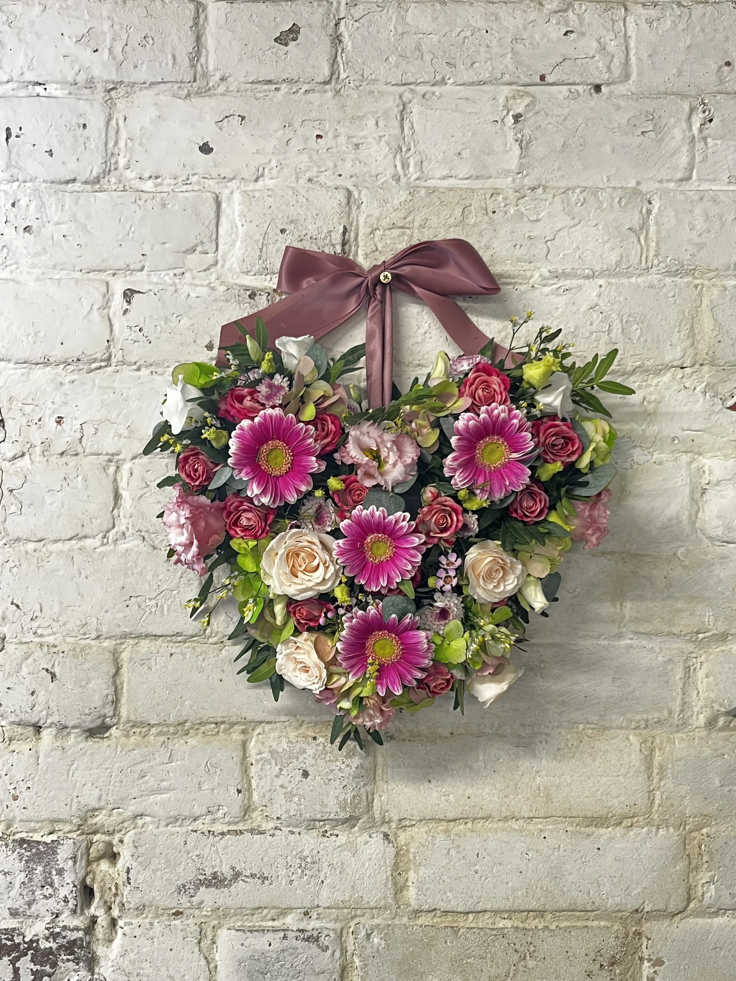 A heart-shaped floral arrangement with pink, white, and green flowers hanging on a white brick wall, decorated with a pinkish-brown bow at the top.