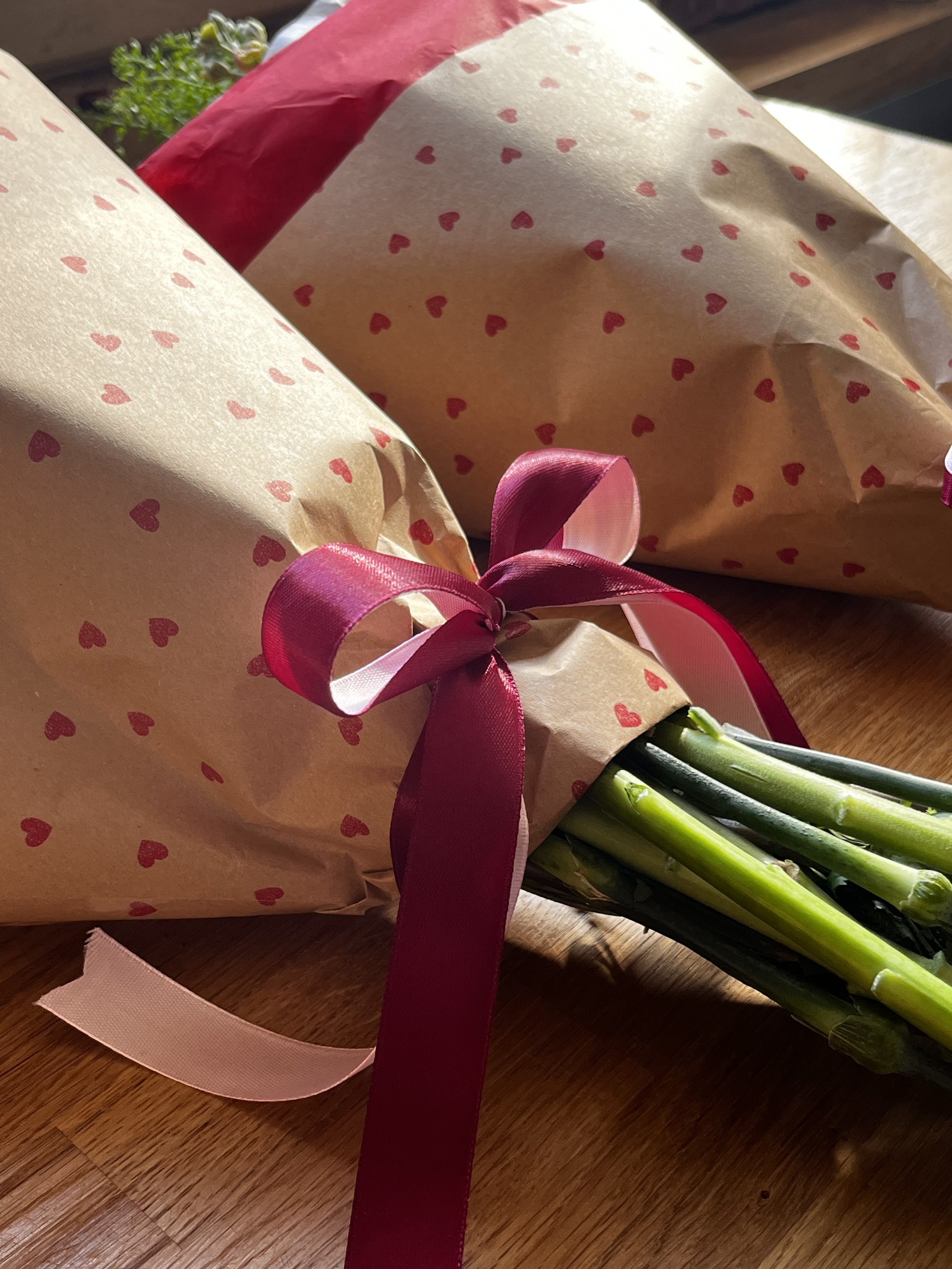 Bouquet of asparagus wrapped in pink and beige paper with small red hearts, tied with a pink and dark pink ribbon, resting on a wooden surface.