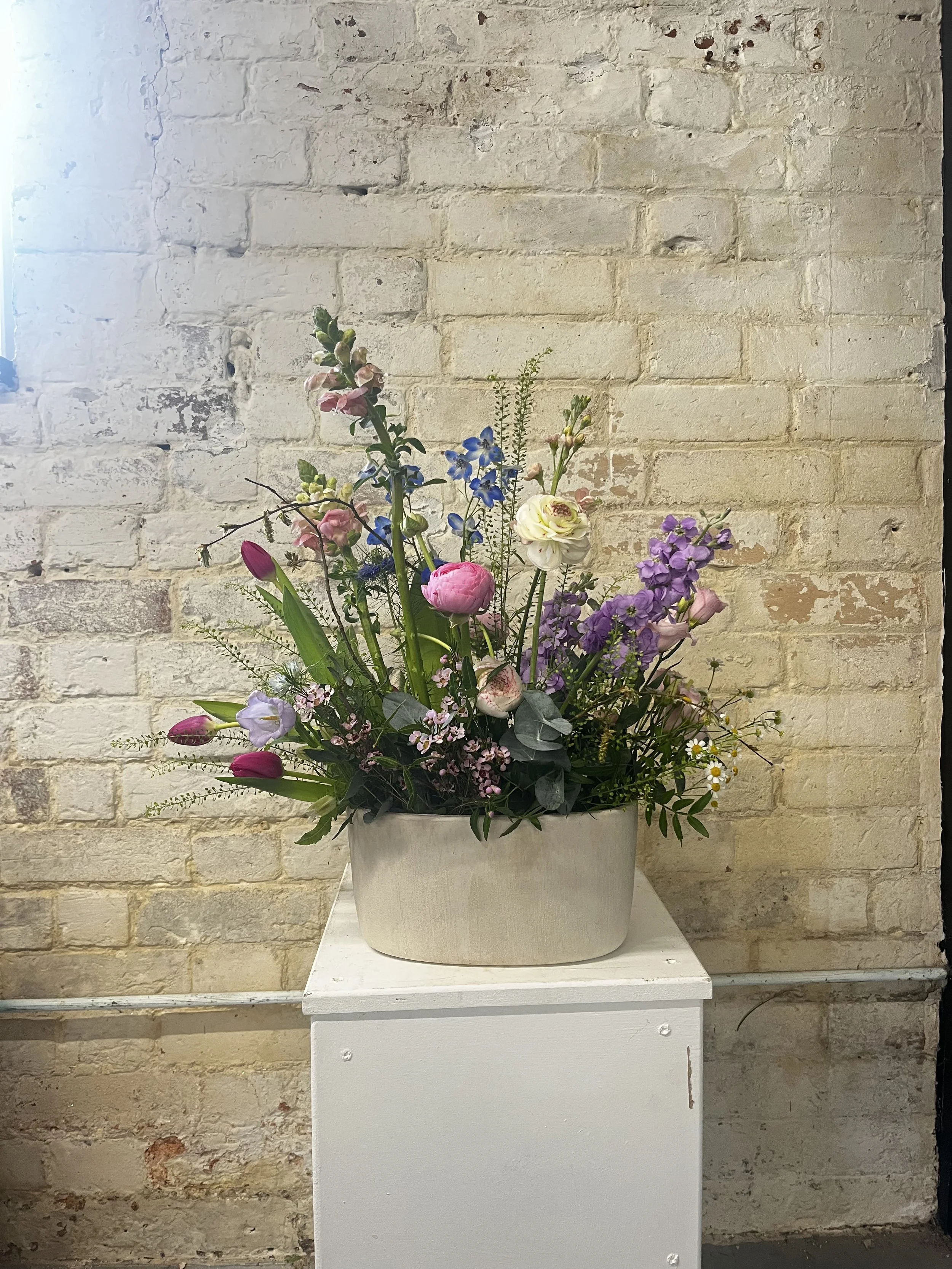 A floral arrangement in a gray ceramic vase on a white pedestal against a exposed brick wall background.