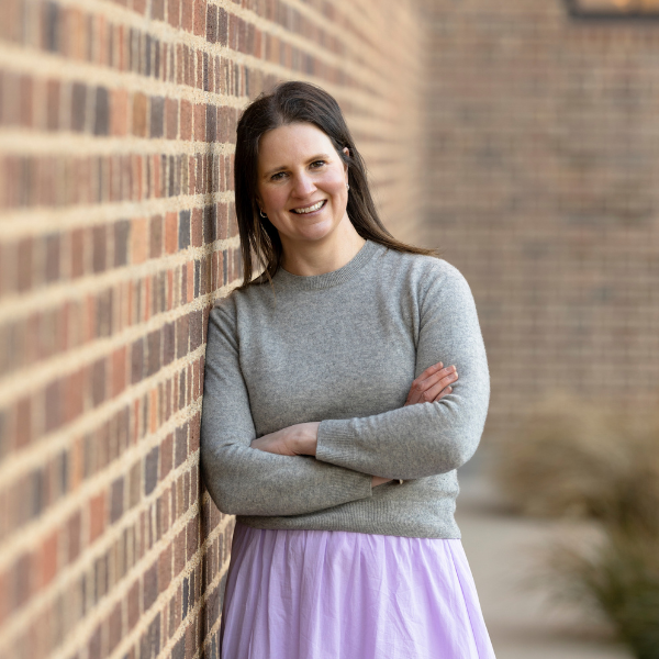 Image of woman wearing a grey jumper and a lilac skirt leaning against a brick wall. Woman has brown hair and is smiling.