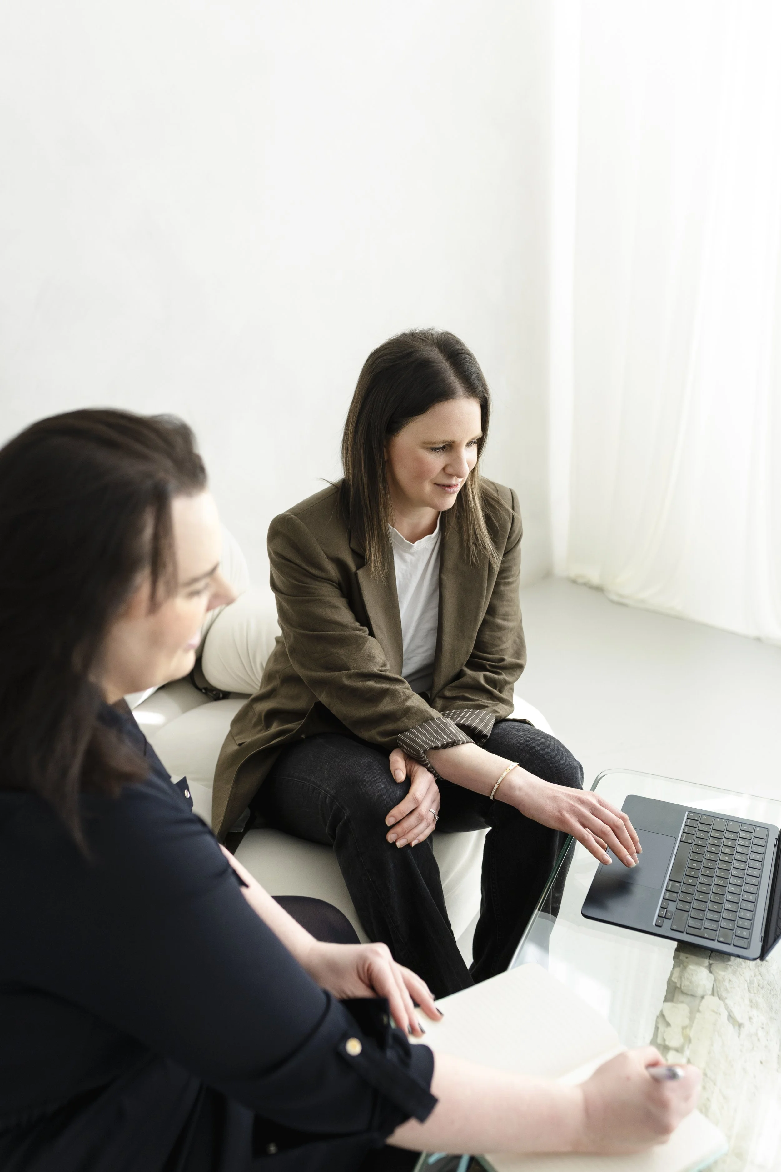 Two females with dark hair sat on a white sofa gathered around a laptop.