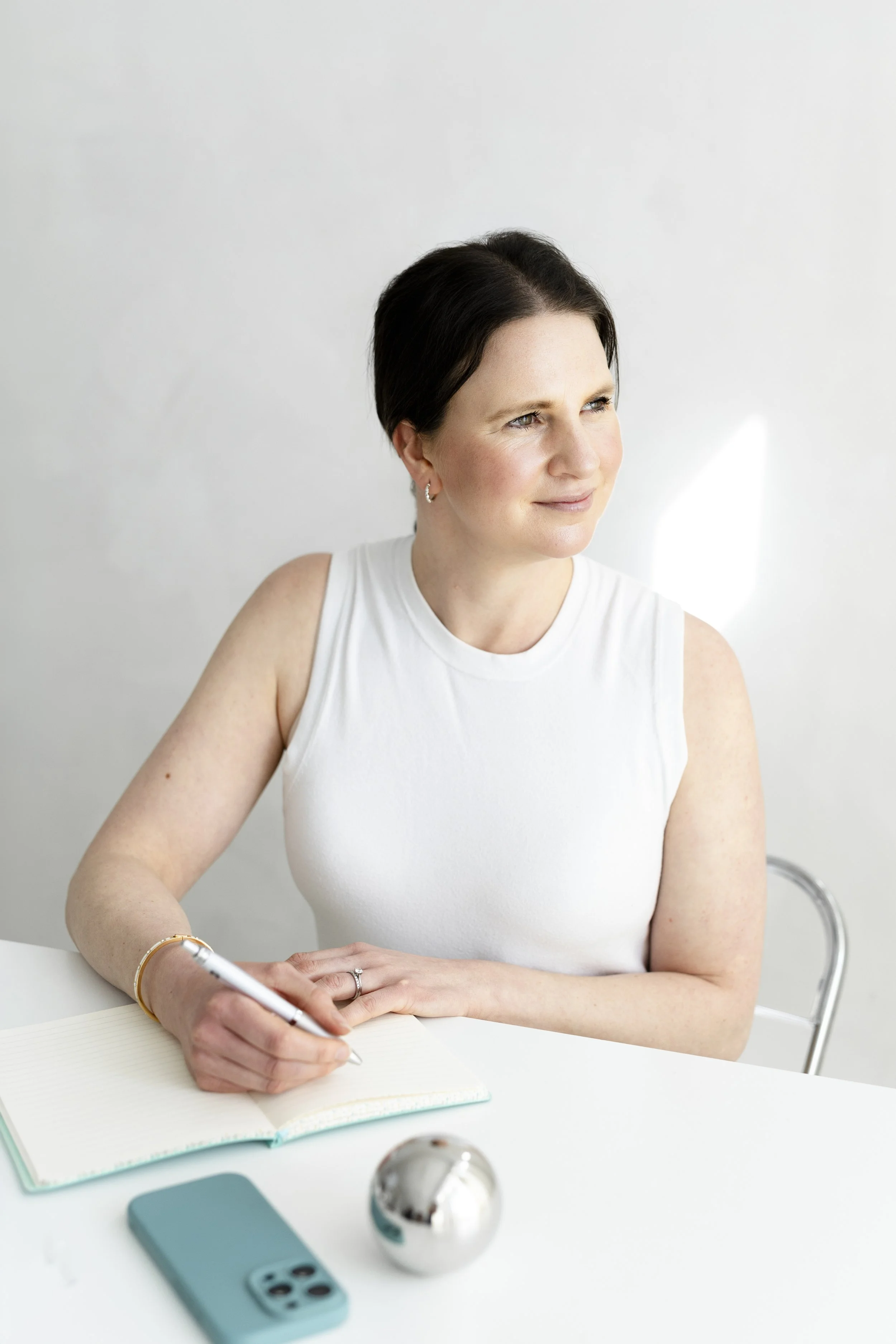Female sat at a white desk on a chair with a silver back, looking into the distance. She has brown hair tied back and is wearing a white sleeveless top. She is writing in a notebook which has a teal cover.
