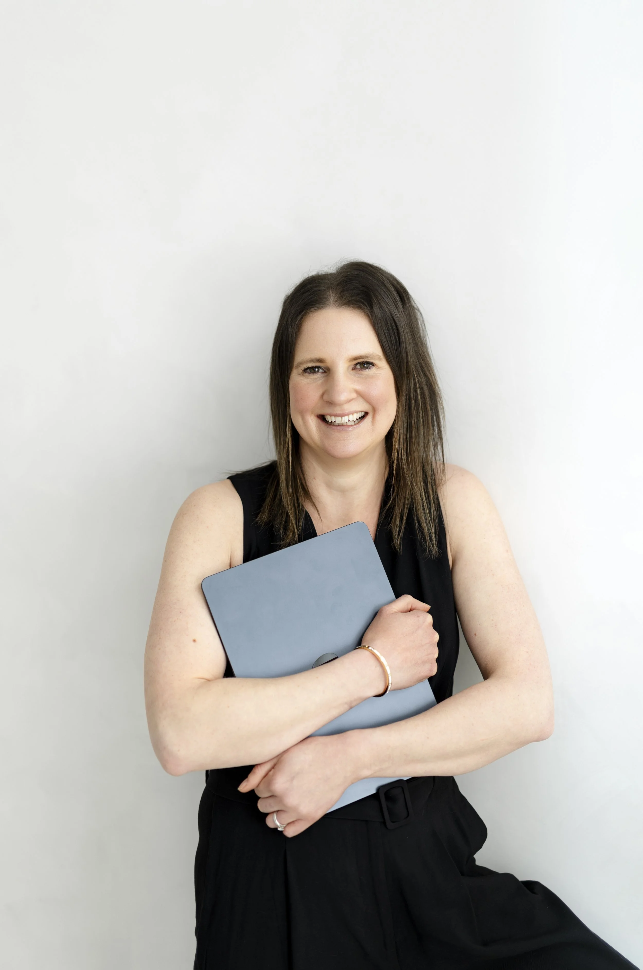 Girl with brown hair and black jumpsuit on sat on stool clutching a dark grey laptop across her chest. She is smiling.