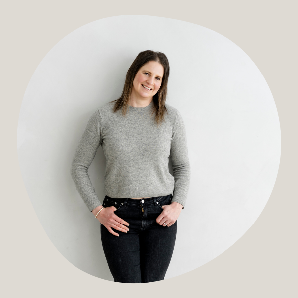 Woman standing up, slightly leaning her head to her left. She is wearing a grey knitted jumper and black jeans. Her thumbs are tucked into each jean pocket.
