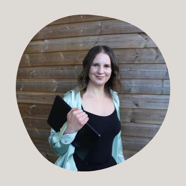 Image of lady standing against wooden shed. She's holding a laptop and wearing a black top and pale green cardigan.