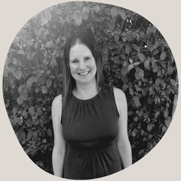 A woman with shoulder-length dark hair, smiling, wearing a sleeveless dark dress, standing in front of a bush with dense leaves.