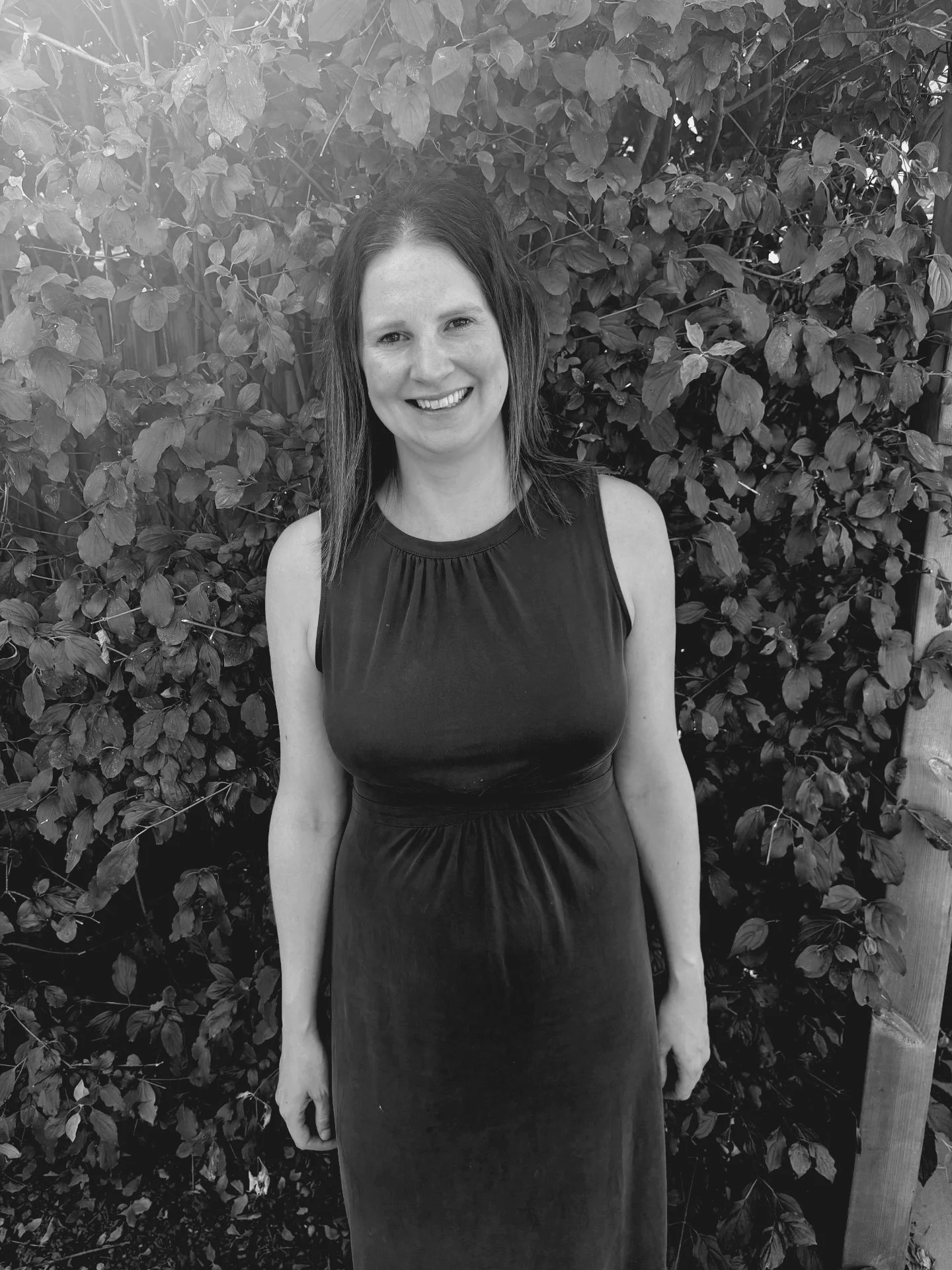 A woman with shoulder-length dark hair, wearing a sleeveless black dress, standing outdoors in front of a leafy shrub, smiling at the camera.