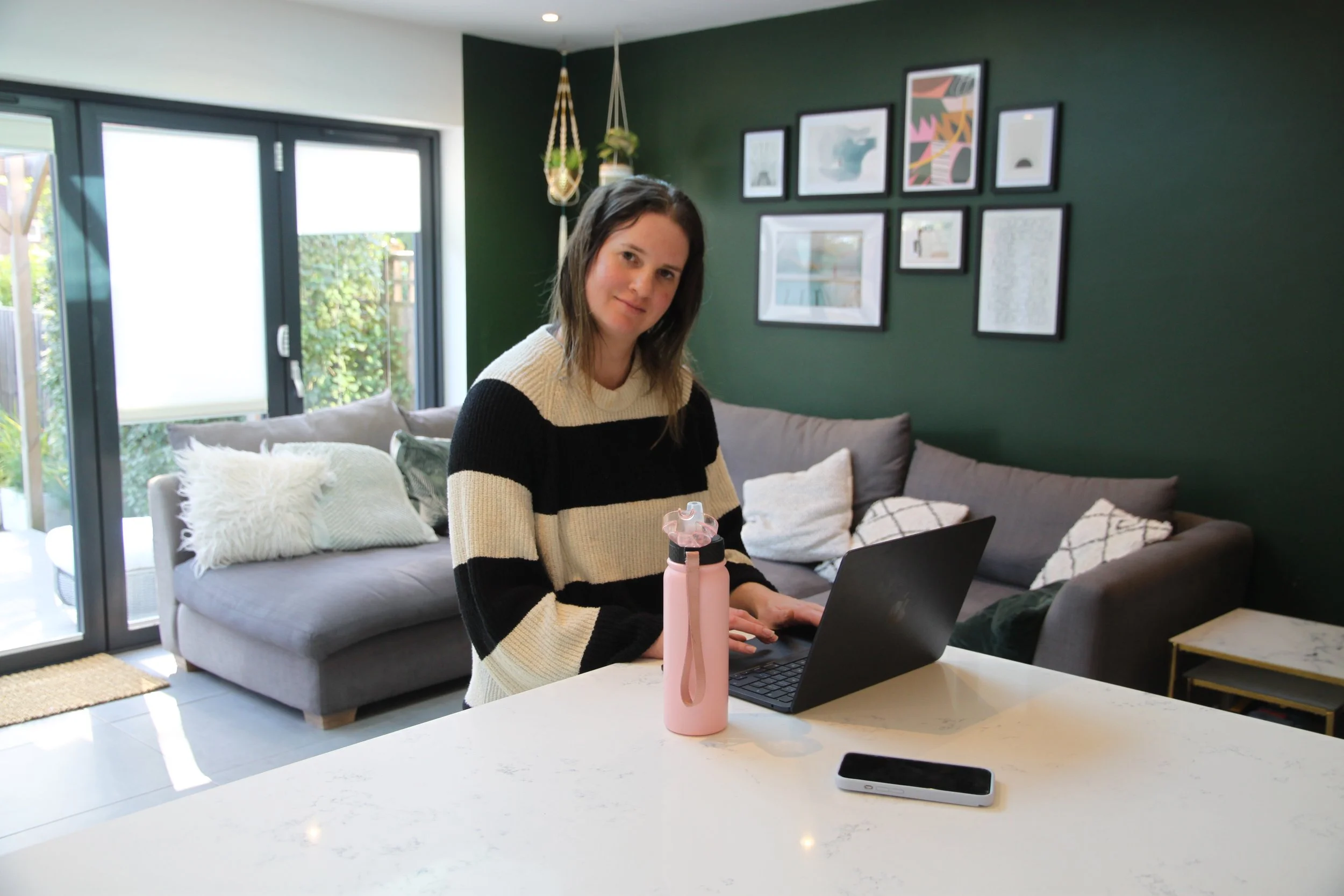 Woman sitting at a kitchen counter with laptop, water bottle, and smartphone in a modern living room with gallery wall and large window.