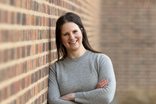 Lady with brown shoulder length hair smiling. She has a grey jumper on and is leaning against a red/brown brick wall.