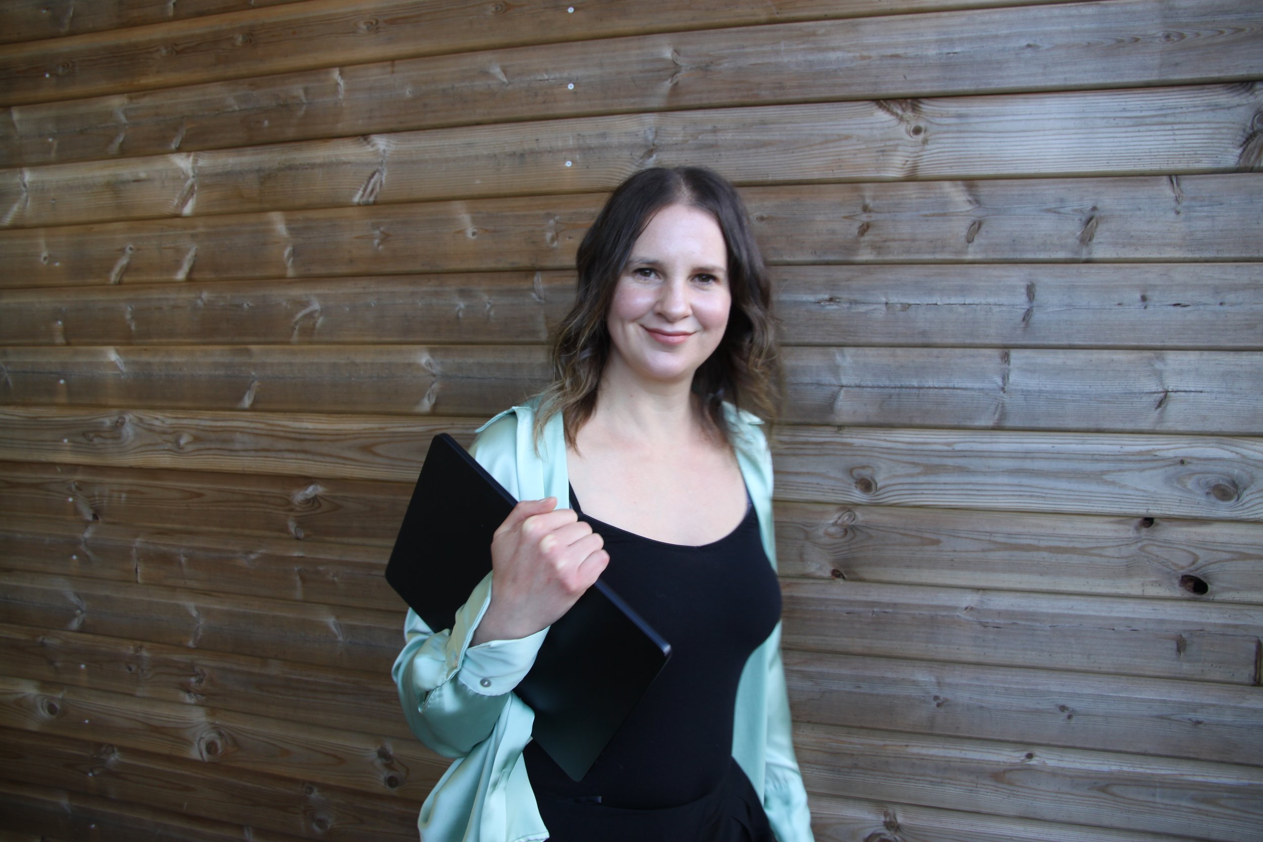 A woman with shoulder-length brown hair, wearing a black top and a light-colored jacket, holding a black laptop, standing against a wooden wall and smiling.