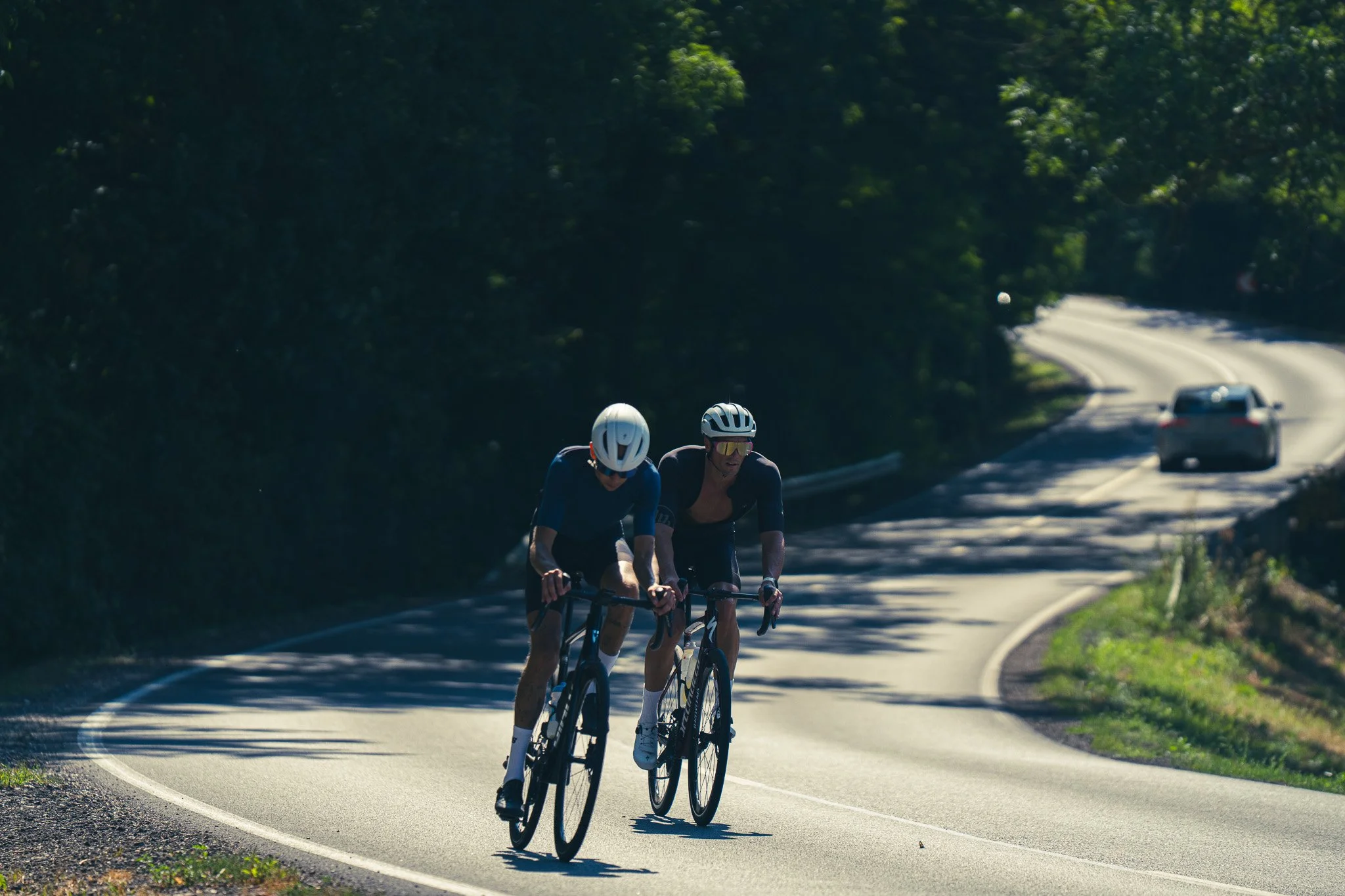 Two cyclists wearing helmets and sunglasses riding on a winding road through a wooded area with a car following behind.