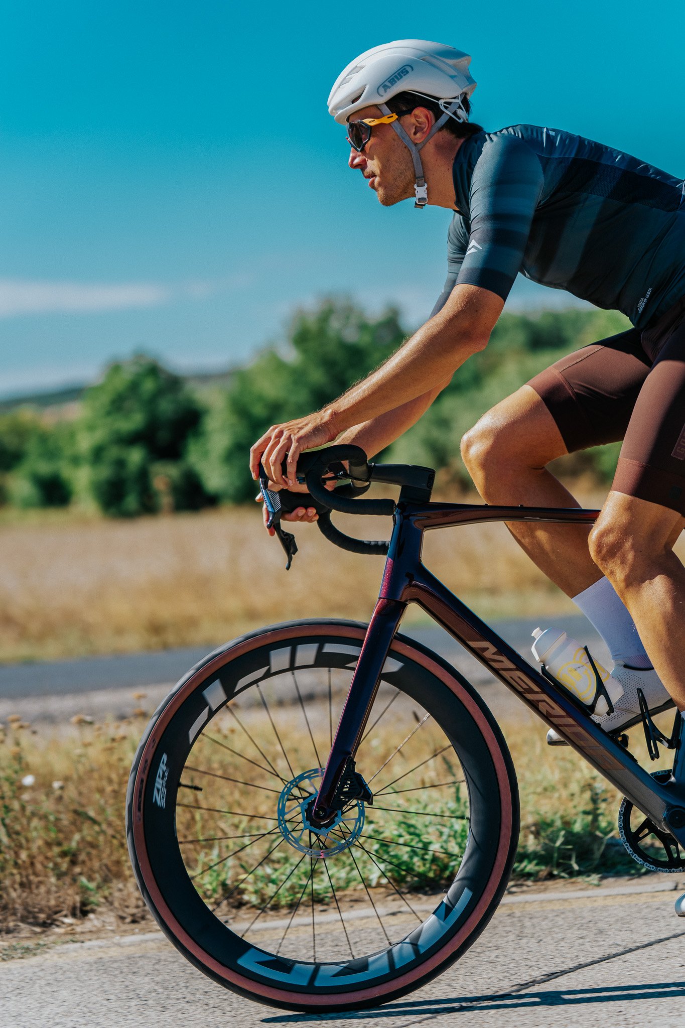 A cyclist riding on a paved road wearing a white helmet, sunglasses, and a dark athletic uniform, with a background of clear blue sky and green trees.