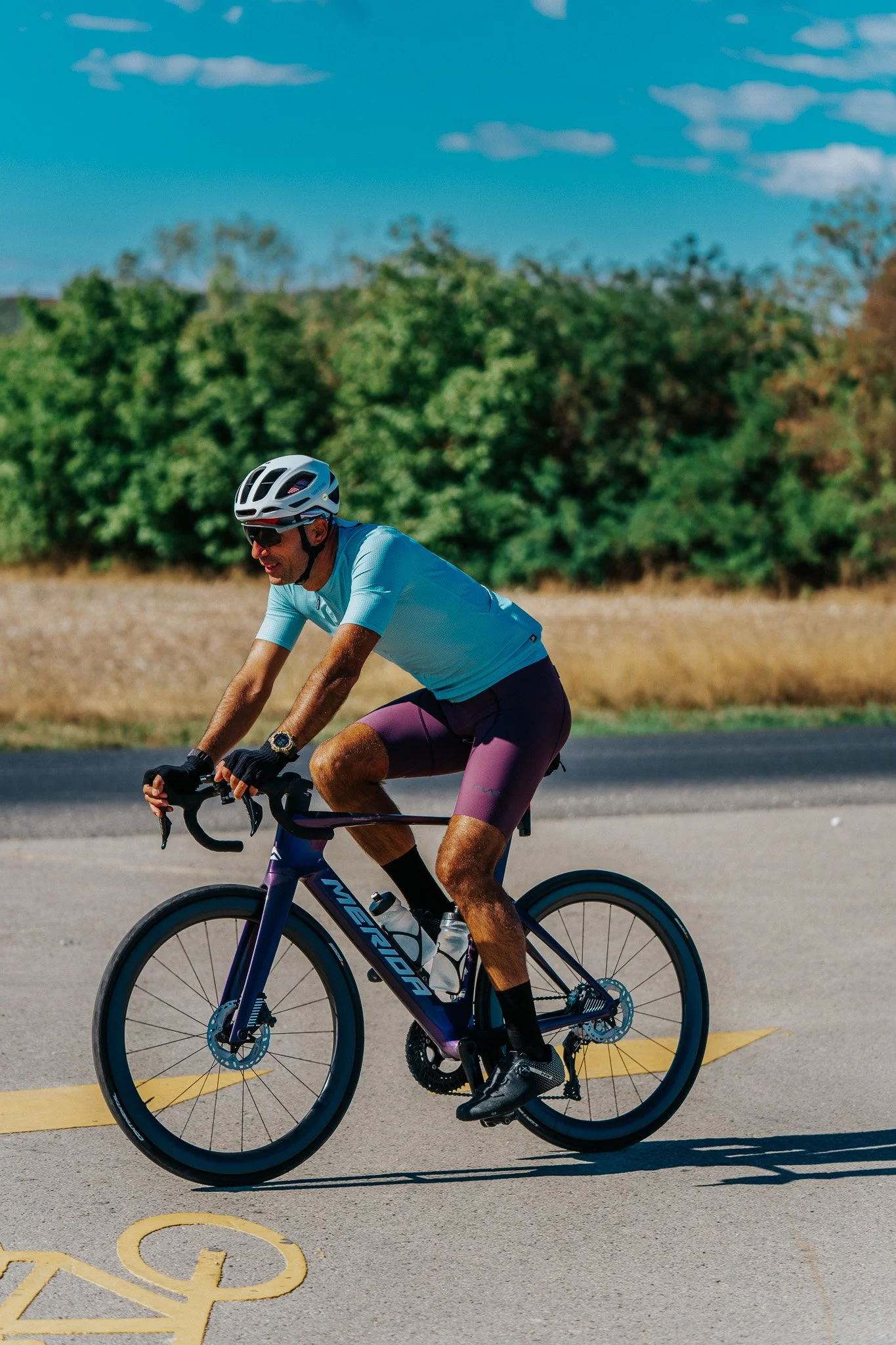 A man wearing a white helmet and sunglasses riding a purple and black road bicycle on a paved road with yellow lane markings, with green trees and a blue sky with clouds in the background.