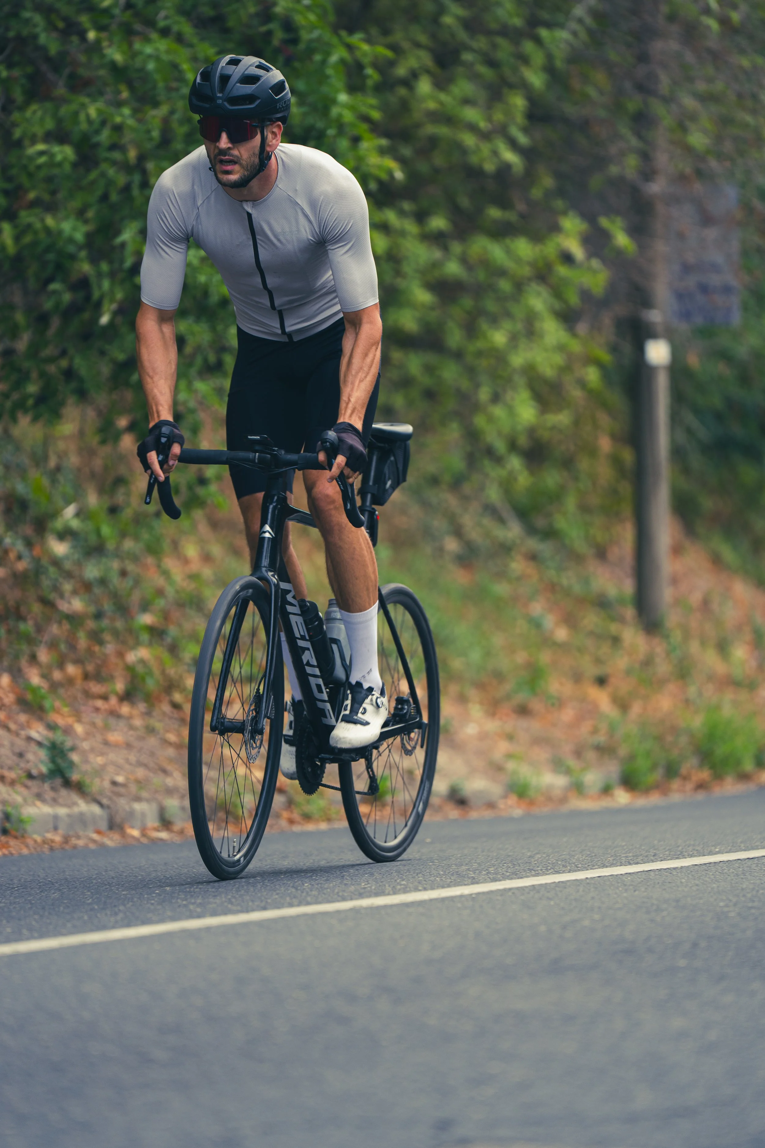 A man riding a black road bicycle on a paved road in a natural outdoor setting. He is wearing a black helmet, sunglasses, a gray cycling jersey, black shorts, and white cycling shoes. Green foliage lines the background.