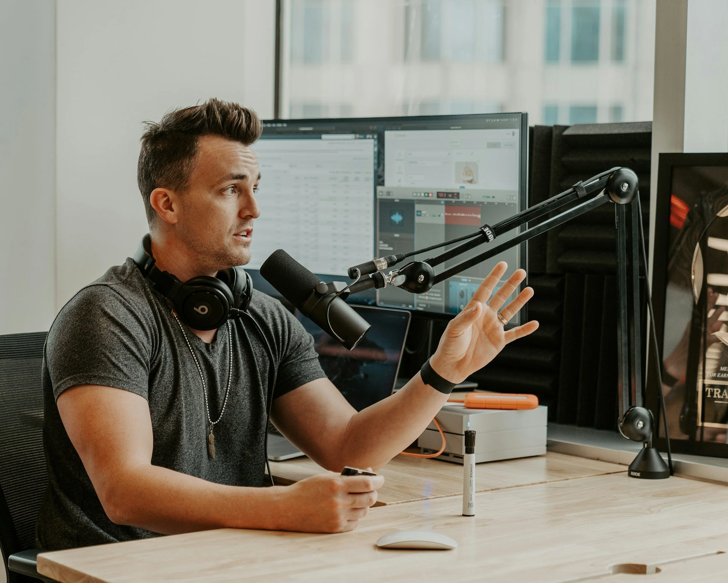 A man with headphones speaking into a microphone in a recording studio with multiple computer monitors in the background.