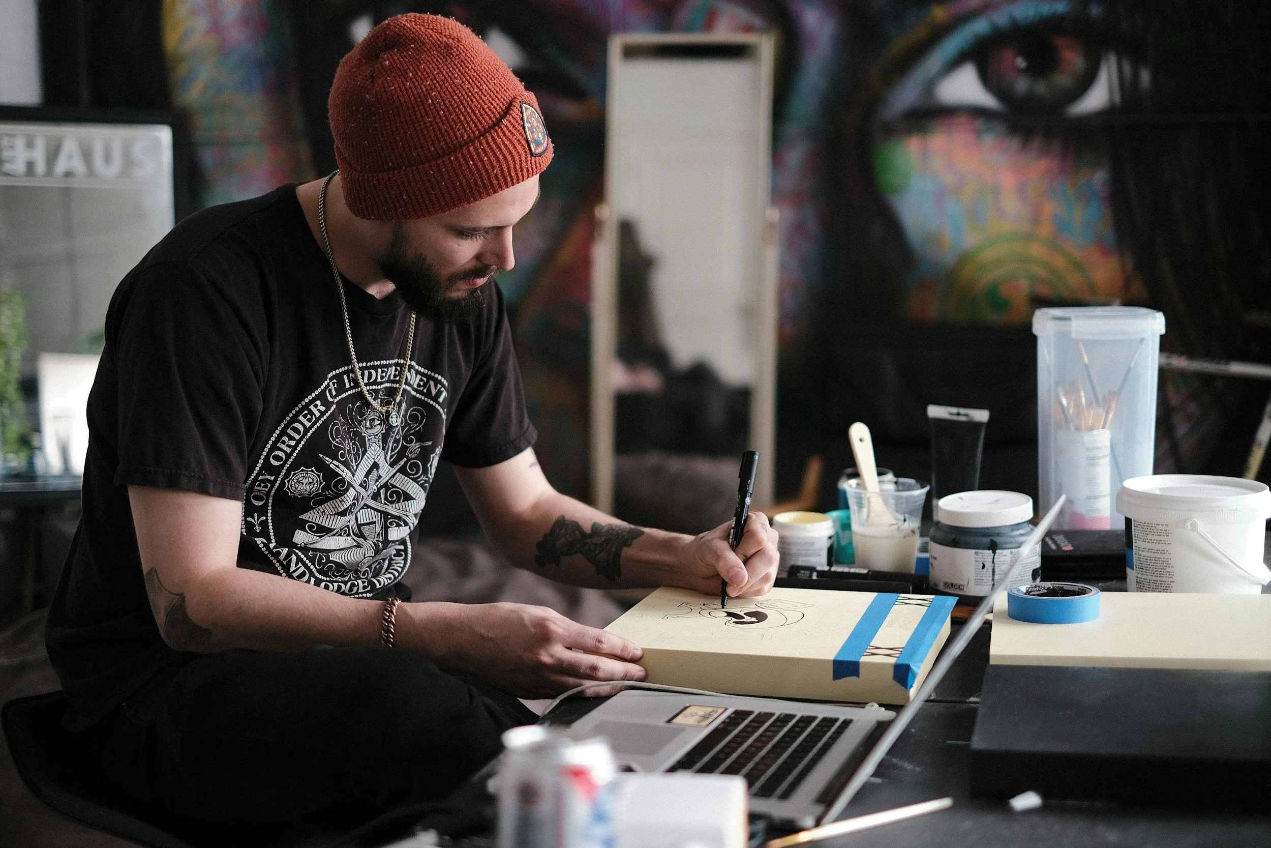 A person with a red beanie and black t-shirt sitting at a table, drawing on a canvas with a black marker. The table has various art supplies, including paint, brushes, and cups. The background features a colorful mural with an eye design.