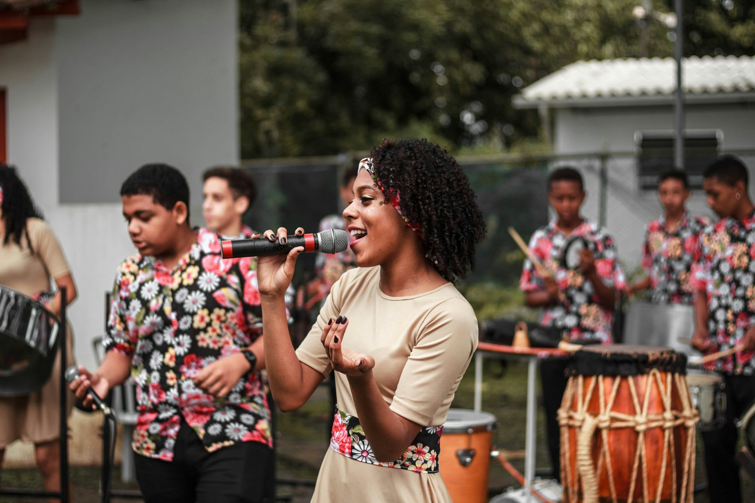 A woman singing into a microphone while a group of children play musical instruments behind her outdoors.