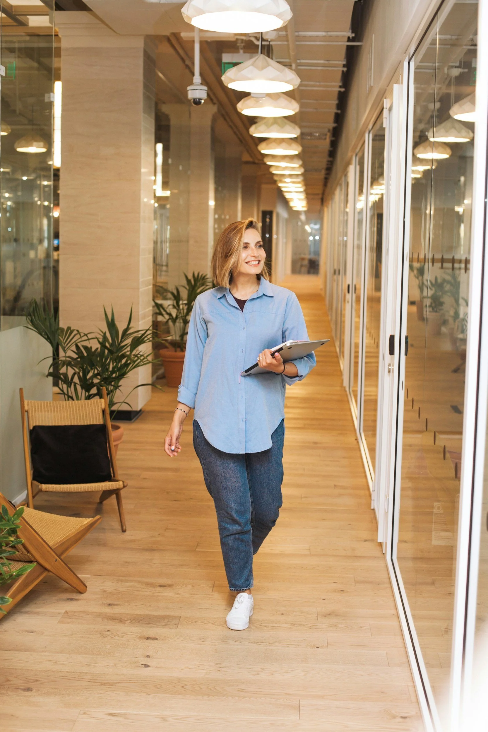 A woman with blonde hair, wearing a light blue shirt and jeans, holding a tablet and walking in a modern office corridor with glass walls and wooden flooring.