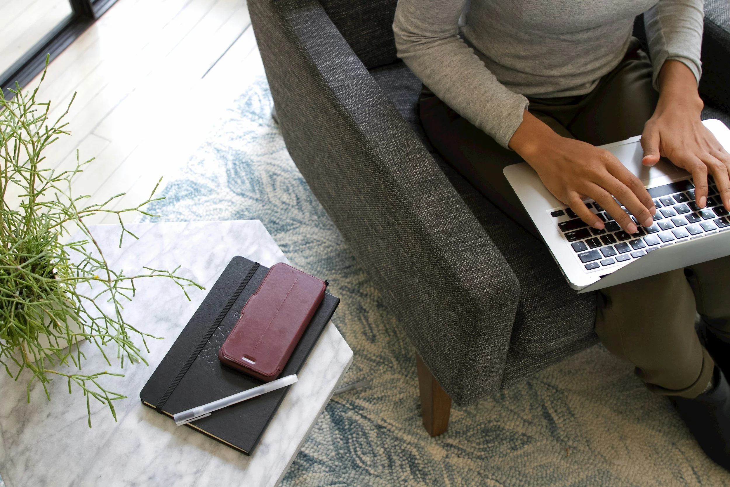 Person sitting on a dark gray sofa using a silver laptop. Nearby on a white marble coffee table are a black notebook, a burgundy phone case, a white pen, and a black pen. There is a green plant on the table, and a patterned rug on the floor.