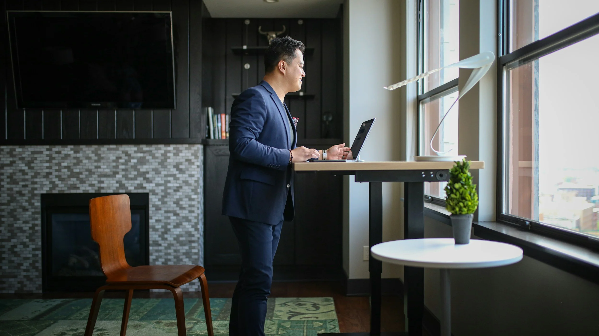 A man in a navy blue suit working on a laptop at a standing desk by large windows in a modern living room.