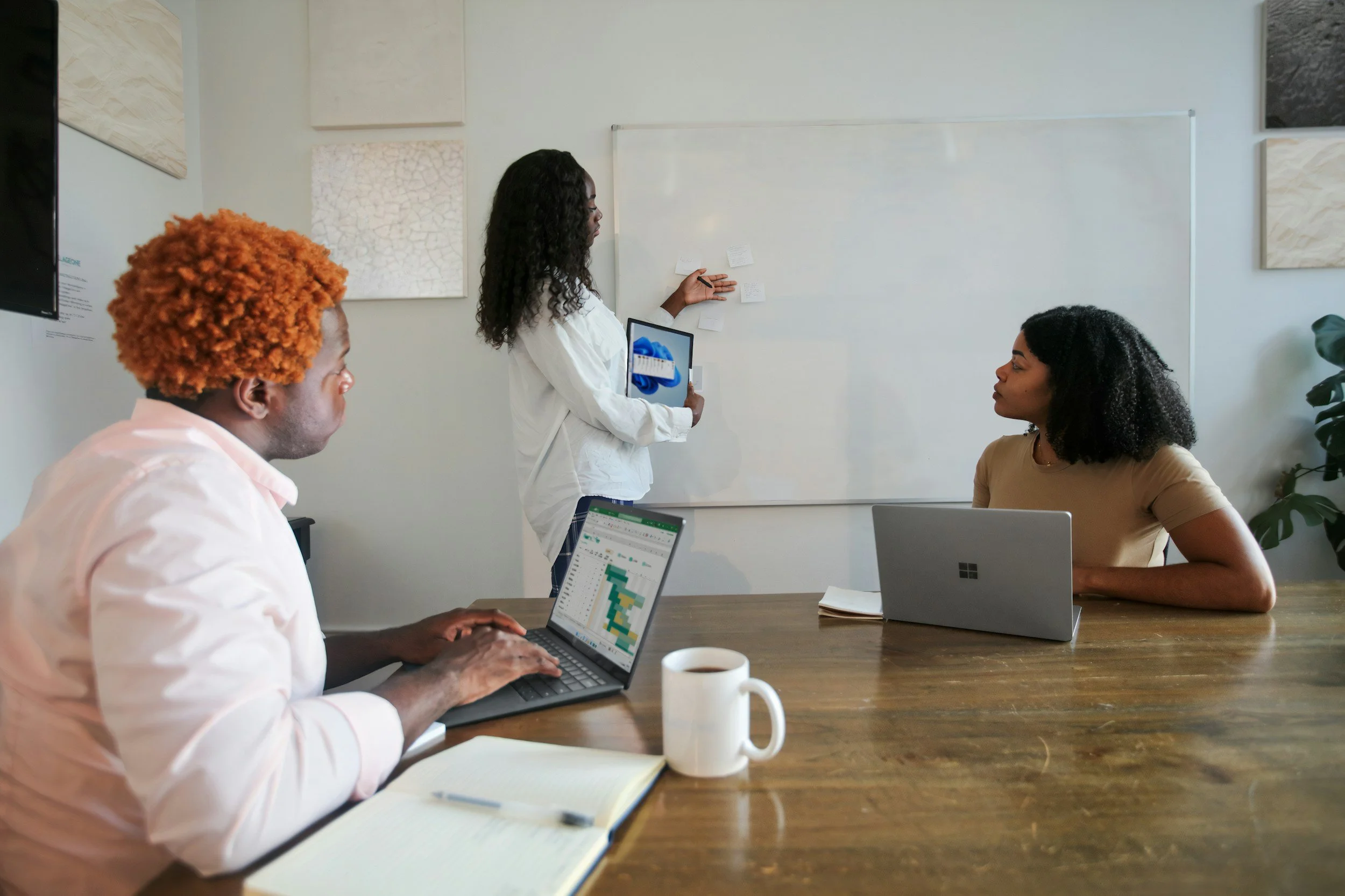 A group of three women in a meeting room, one standing and two seated at a wooden table, with laptops, a notebook, and a coffee mug, while the standing woman presents information on a whiteboard.