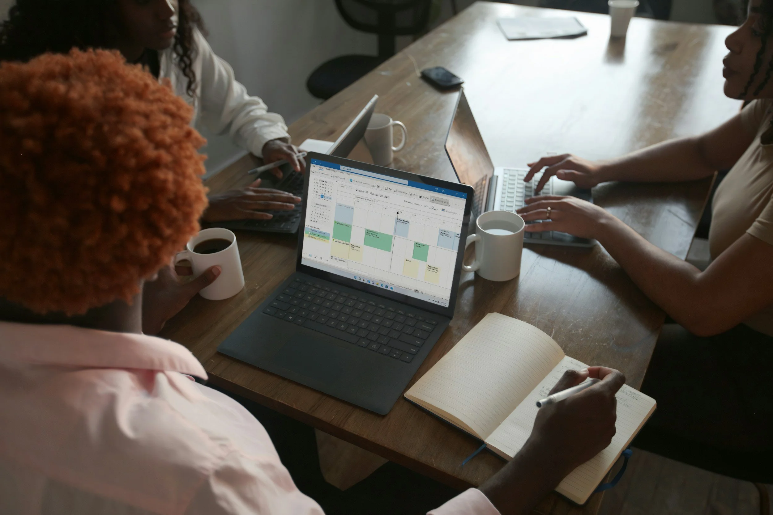 Three women working together at a wooden table with laptops, notebooks, and cups of coffee.