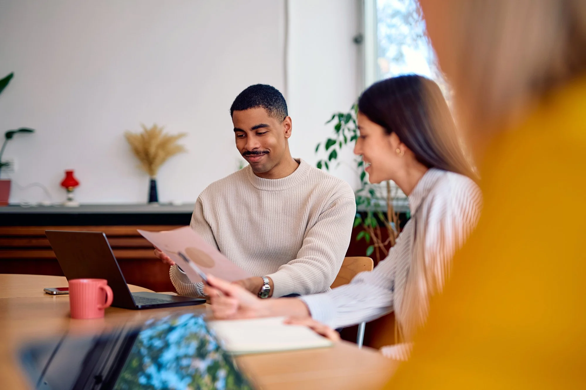 Two people, a man and a woman, sitting at a wooden table in an office or meeting room, reviewing documents together. The man has short black hair and is wearing a beige sweater, while the woman has long dark hair and is wearing a white striped blouse. They are smiling and appear engaged in a discussion, with a pink mug, a laptop, and a smartphone on the table.
