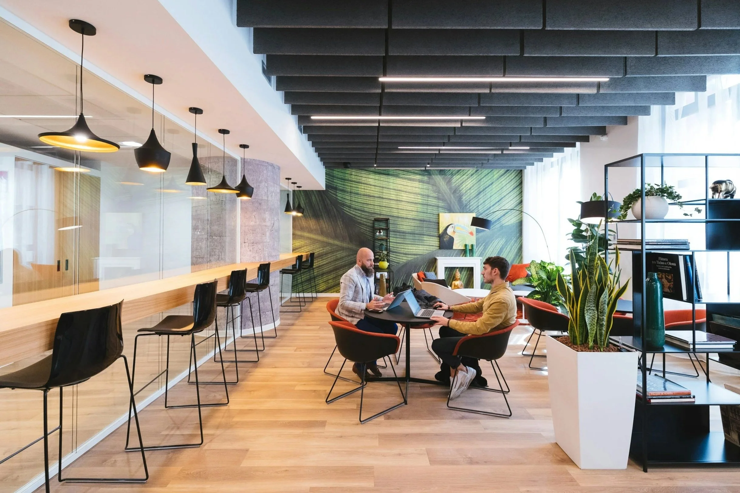 Modern office lounge with two men working on laptops at a round table, surrounded by plants, colorful chairs, and a vibrant green leaf mural on the wall.
