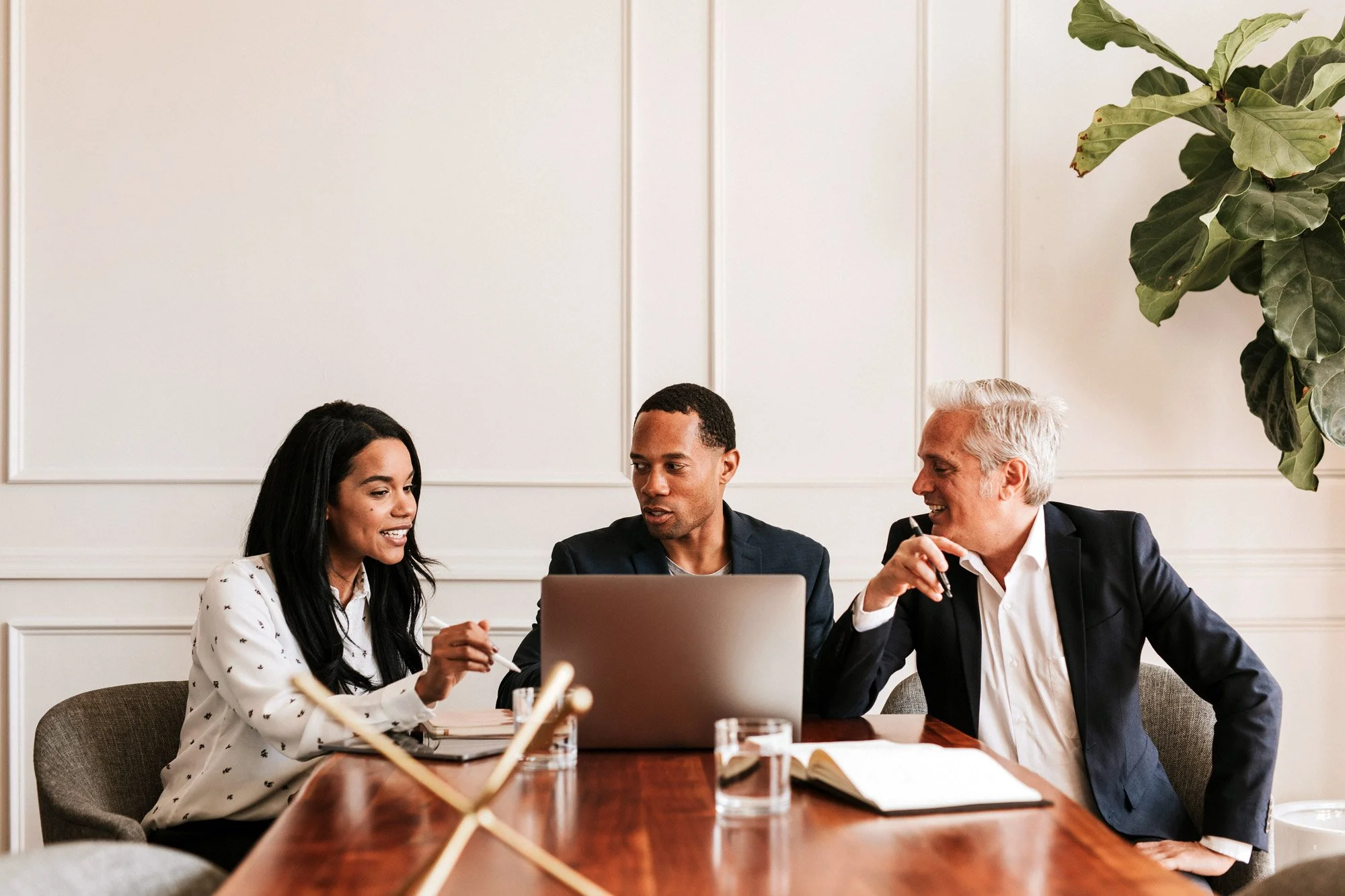 Three professionals, two men and one woman, sitting at a conference table engaged in a discussion in a well-lit office conference room.