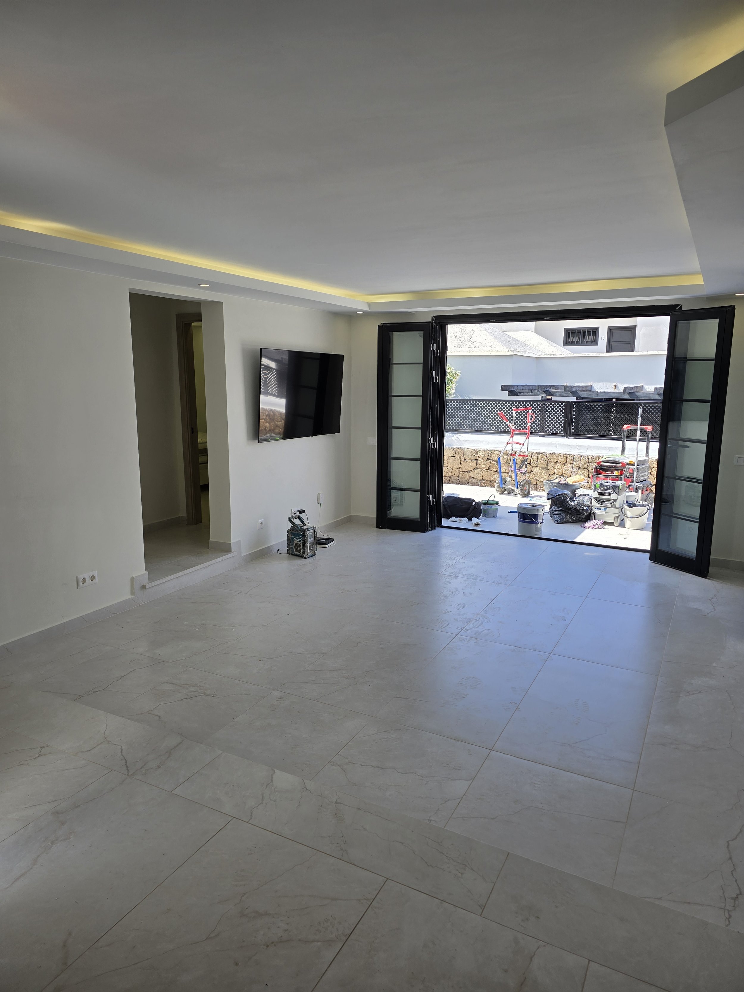 Empty living room with large open glass doors leading to a patio, a wall-mounted TV, and some tools on the floor.