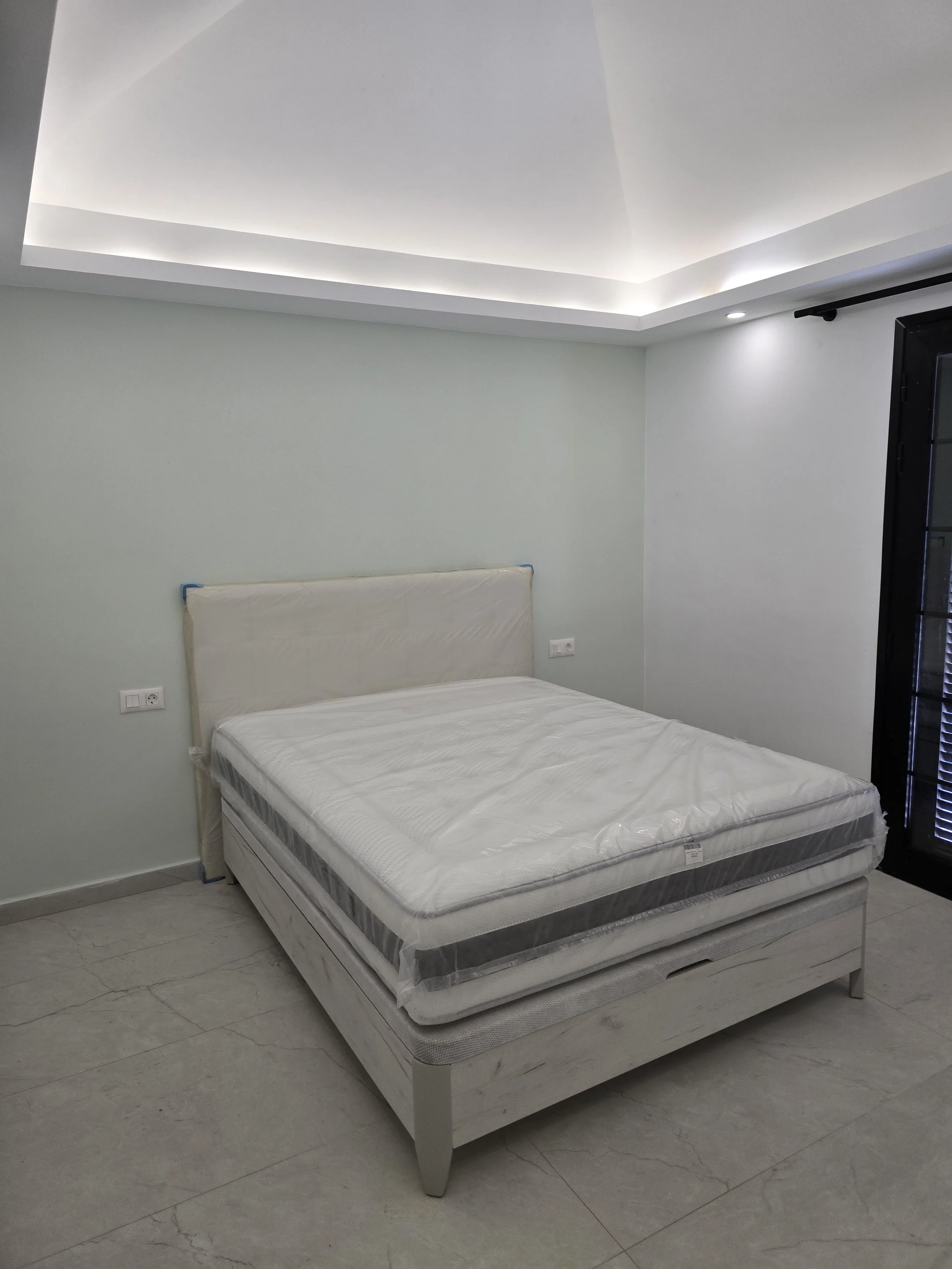 A bedroom with a white bed, new mattress, and light-colored tiled floor. The room has a light green wall, a ceiling with recessed lighting and cove lighting, and a black door with glass panels.
