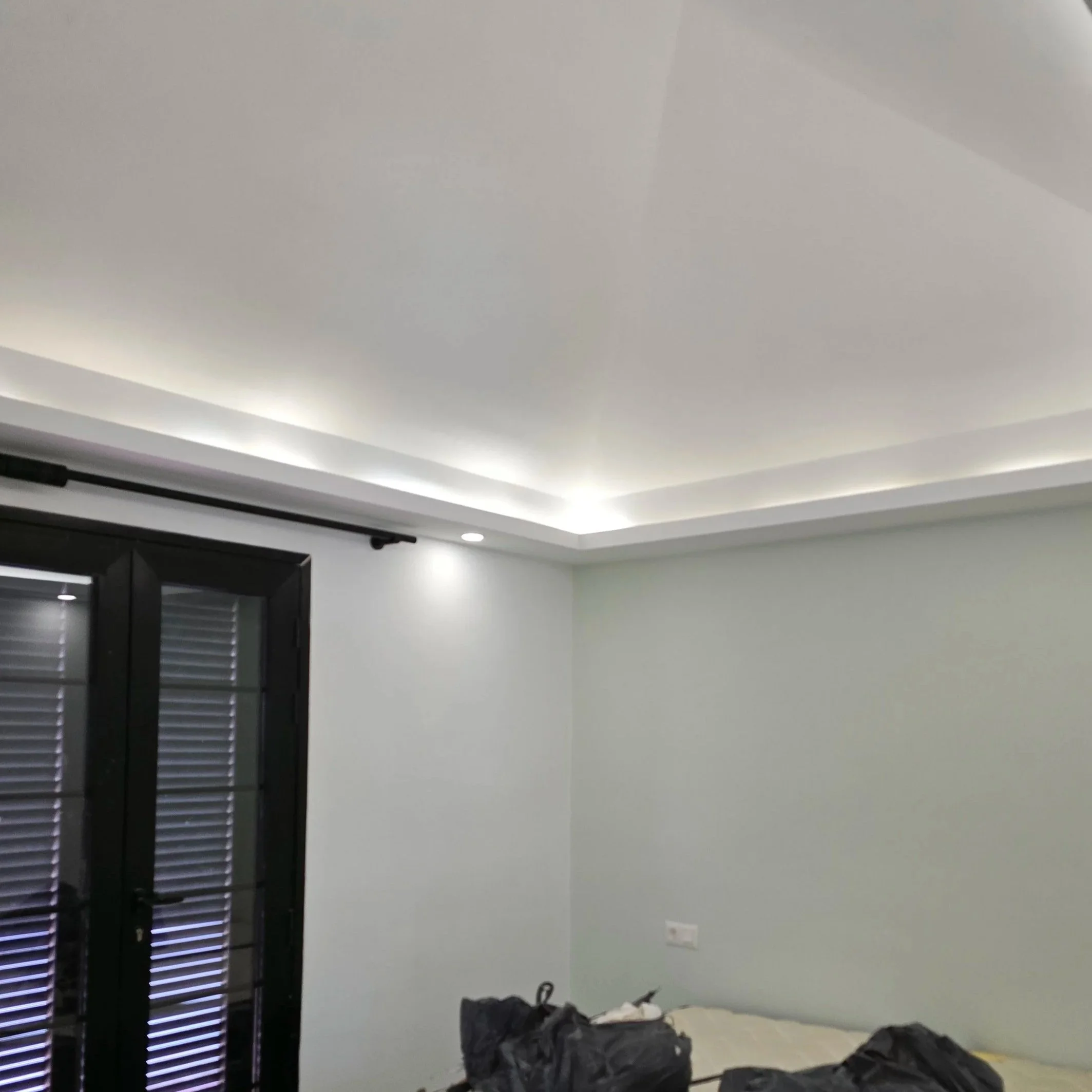 Interior of a modern bedroom with white walls, a ceiling with cove lighting, a black framed door with shutters, and some bags or clothing on the bed.