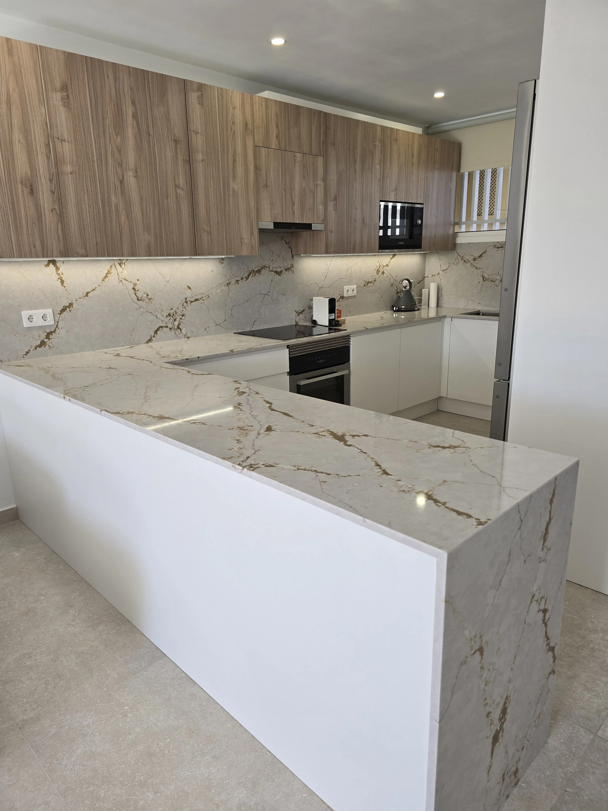 Modern kitchen with marble countertops, wooden upper cabinets, and white lower cabinets. A built-in oven, microwave, and some small appliances are visible.