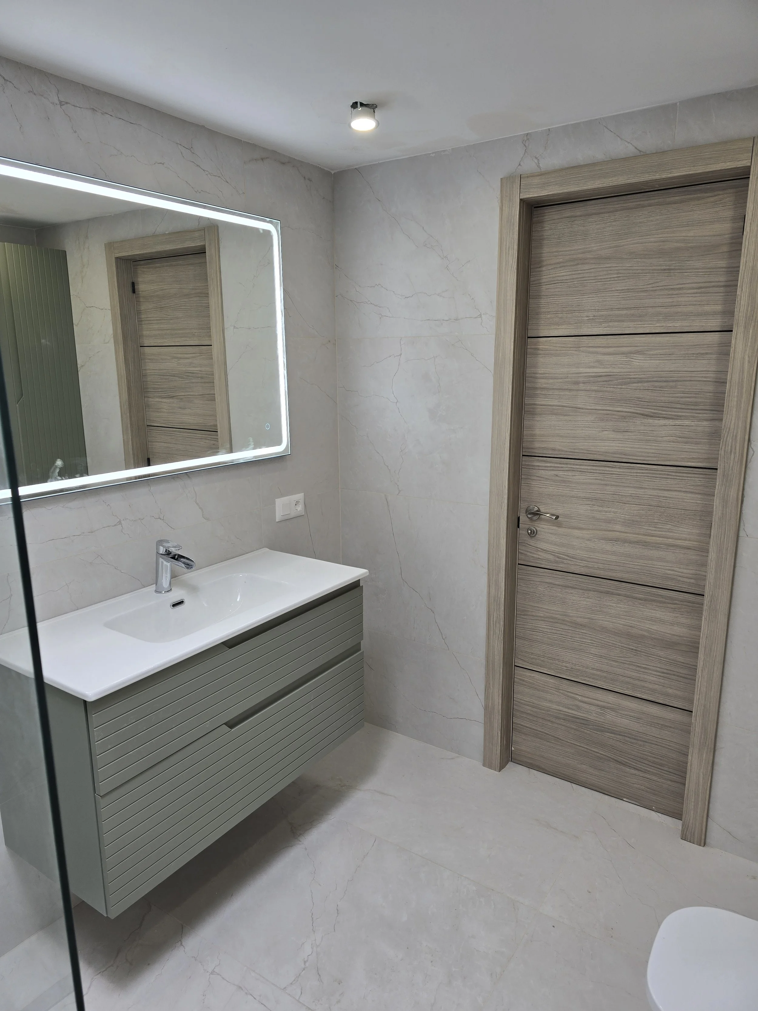 Modern bathroom with beige marble walls and floor, a floating vanity with a white sink and a mirror with LED lighting, a wooden door, and a ceiling light.