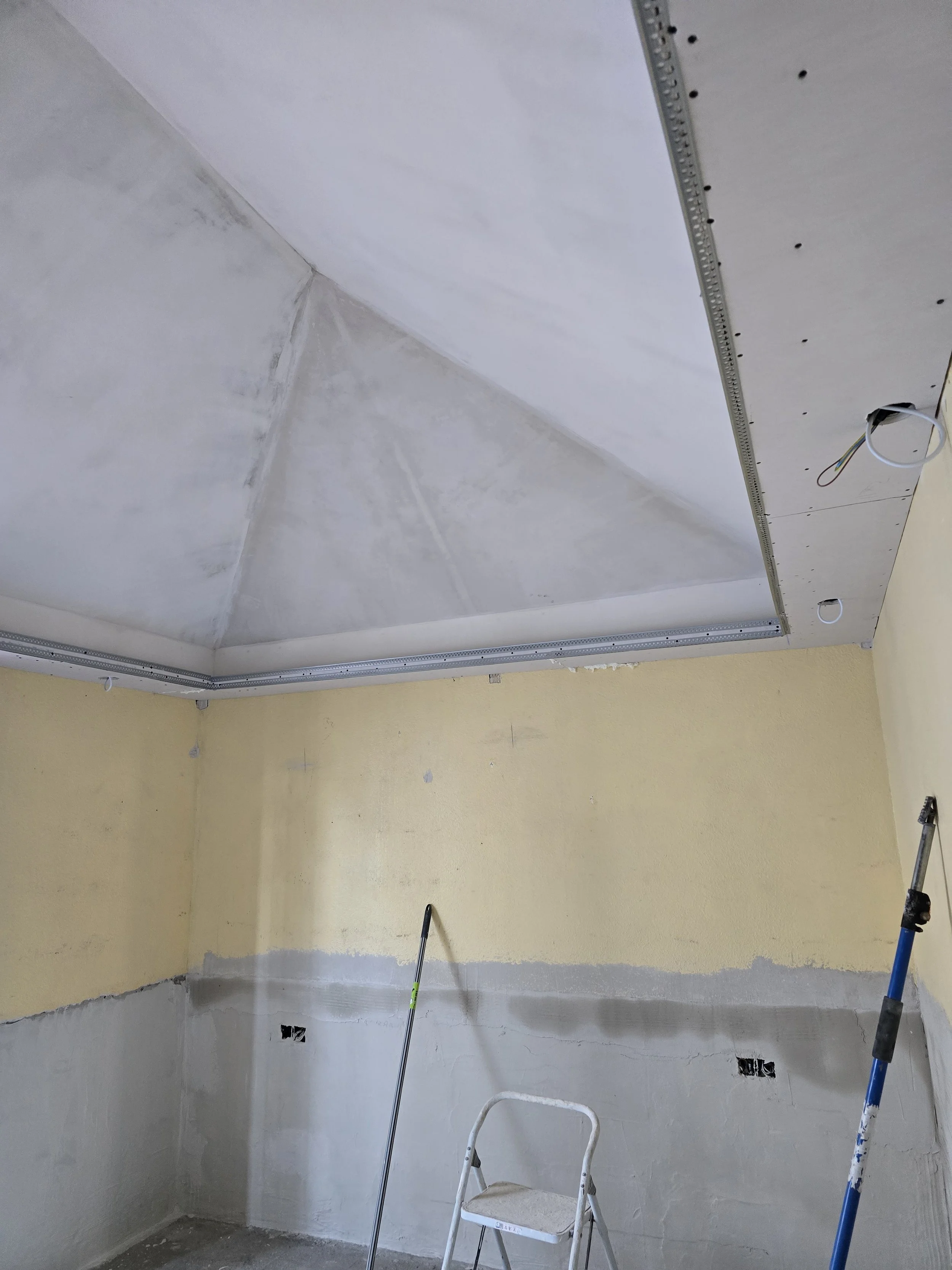 Room under construction with drywall installation on ceiling and walls, ladder, and construction tools.