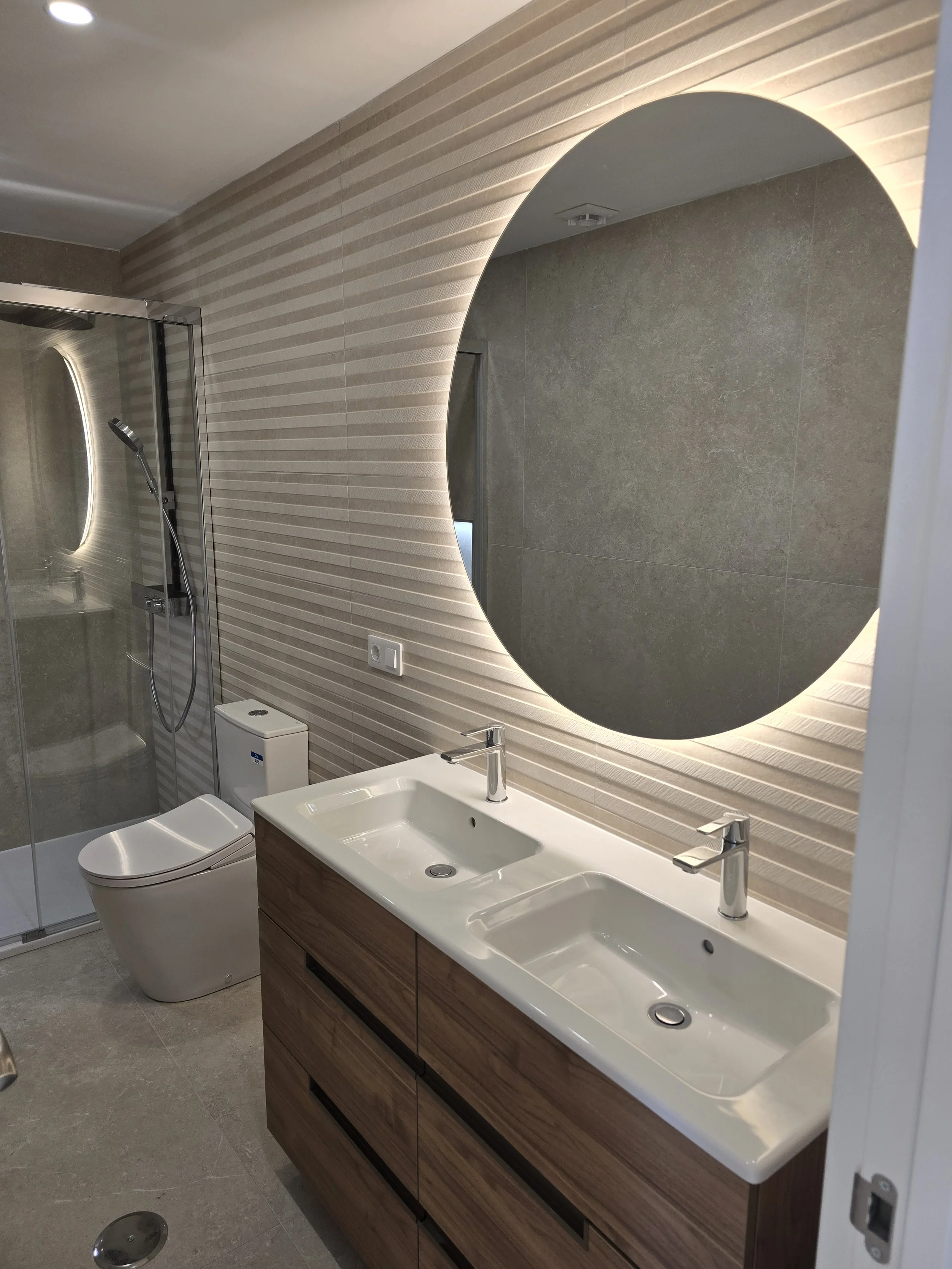 Modern bathroom with dual sinks, large round mirror with backlit lighting, toilet, and a walk-in shower with glass door and handheld showerhead.