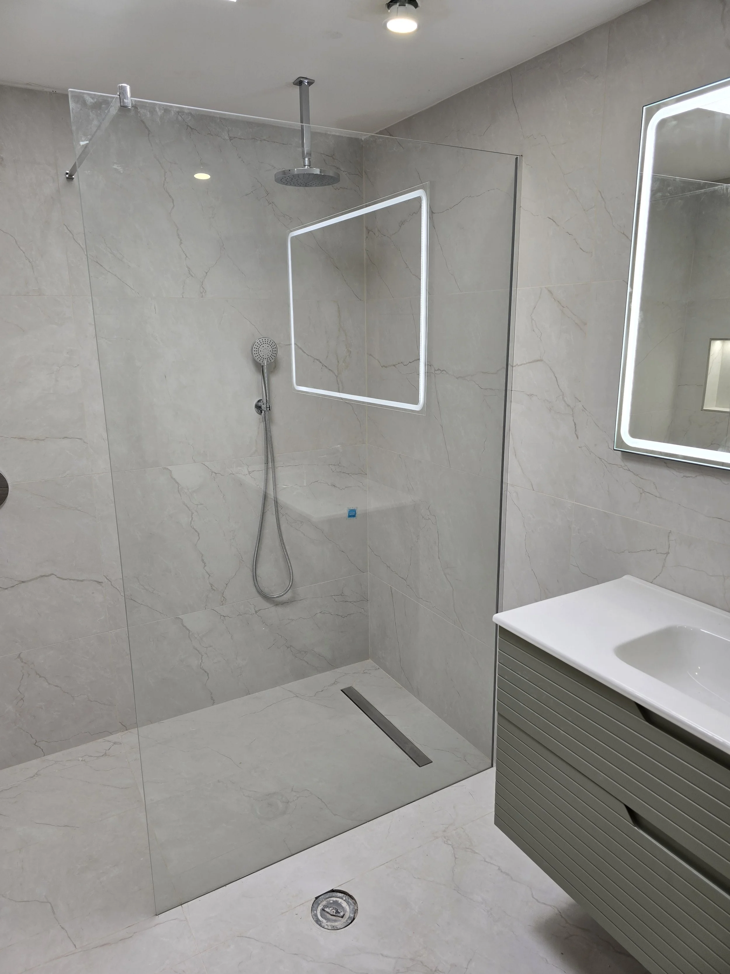 Modern bathroom with a walk-in shower featuring a glass partition, ceiling-mounted rain showerhead, handheld shower, and a large mirror next to a white vanity with a built-in sink.
