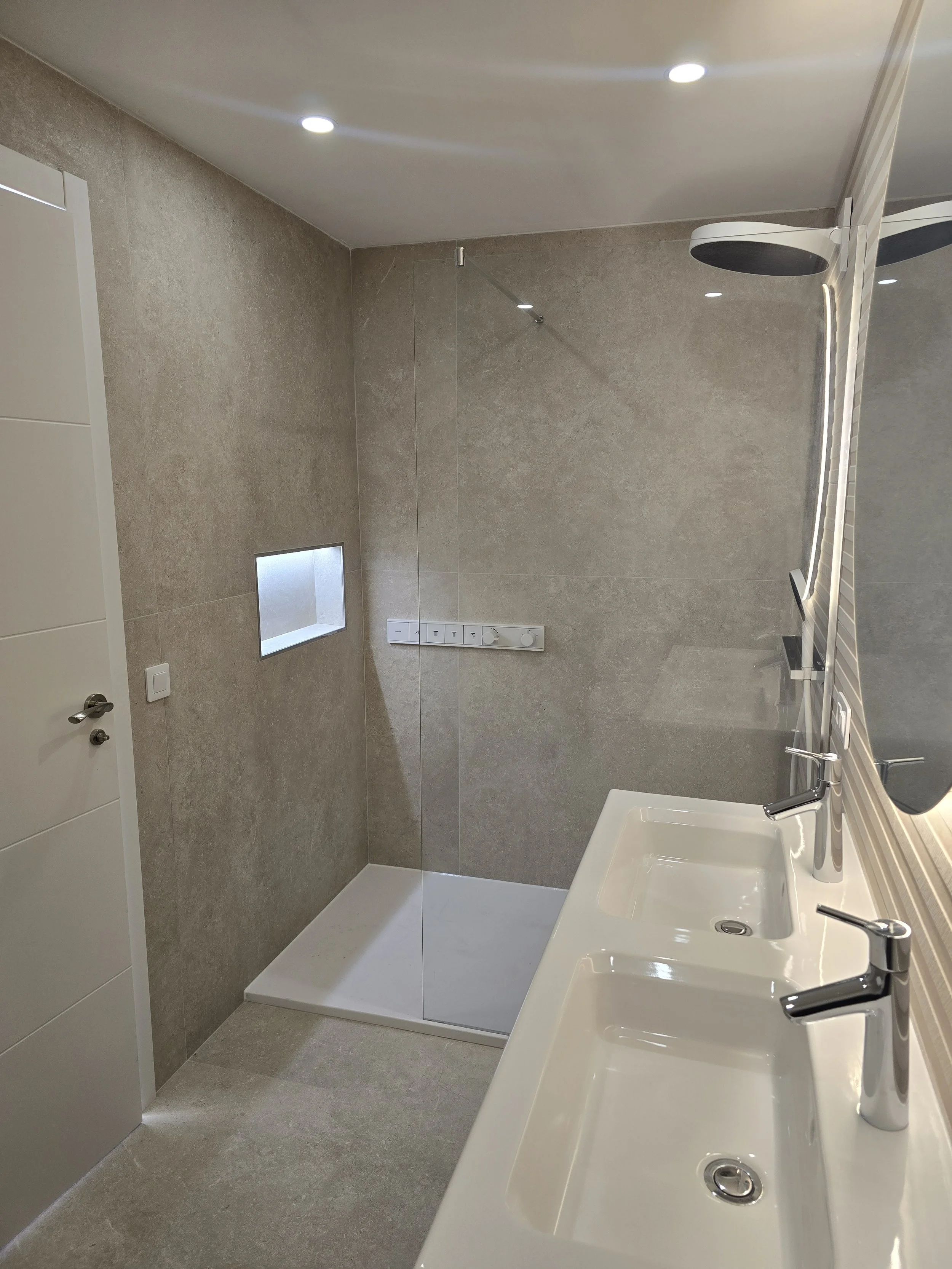 A modern bathroom with a shower area enclosed by a glass panel, a small built-in wall niche in the shower, a double sink vanity with chrome faucets, a large mirror, ceiling lights, and beige tiled walls and floor.