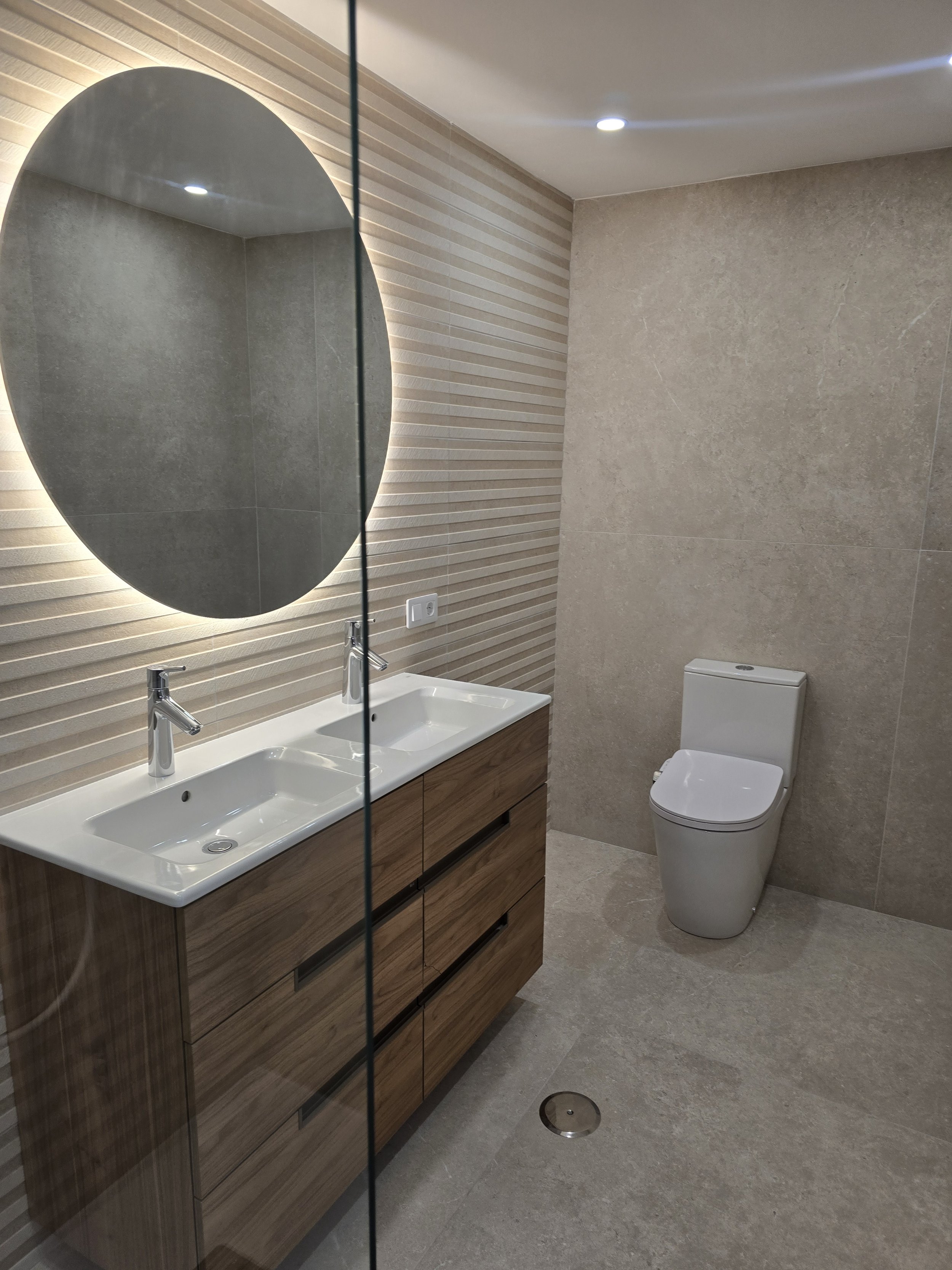 Modern bathroom with dual sinks, a large mirror, and a toilet, featuring beige textured wall tiles and wooden vanity cabinets.
