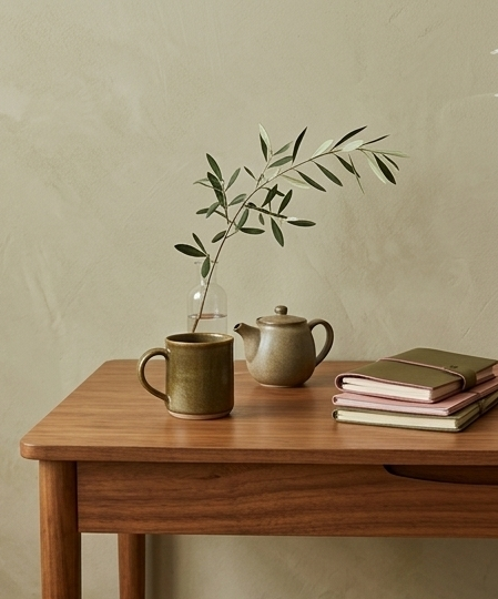 A wooden table holding a green ceramic mug, a beige teapot, and a glass vase with green leafy branches, with several closed books stacked on the right side.