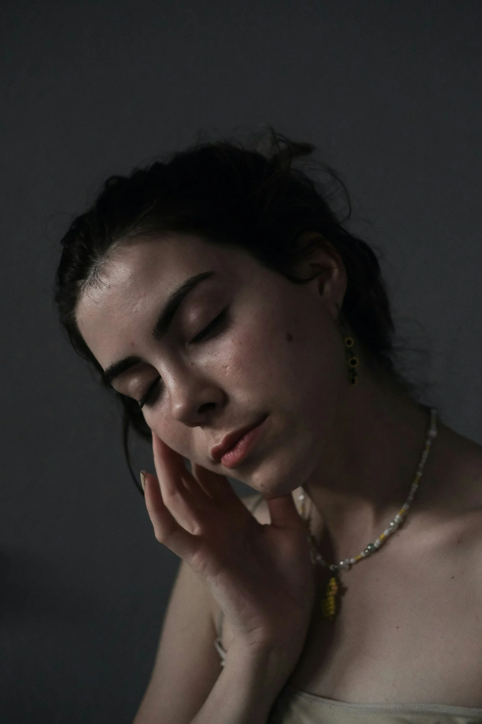 A woman with dark hair, closed eyes, and a peaceful expression, her hand gently resting on her face, wearing a beaded necklace against a dark background.