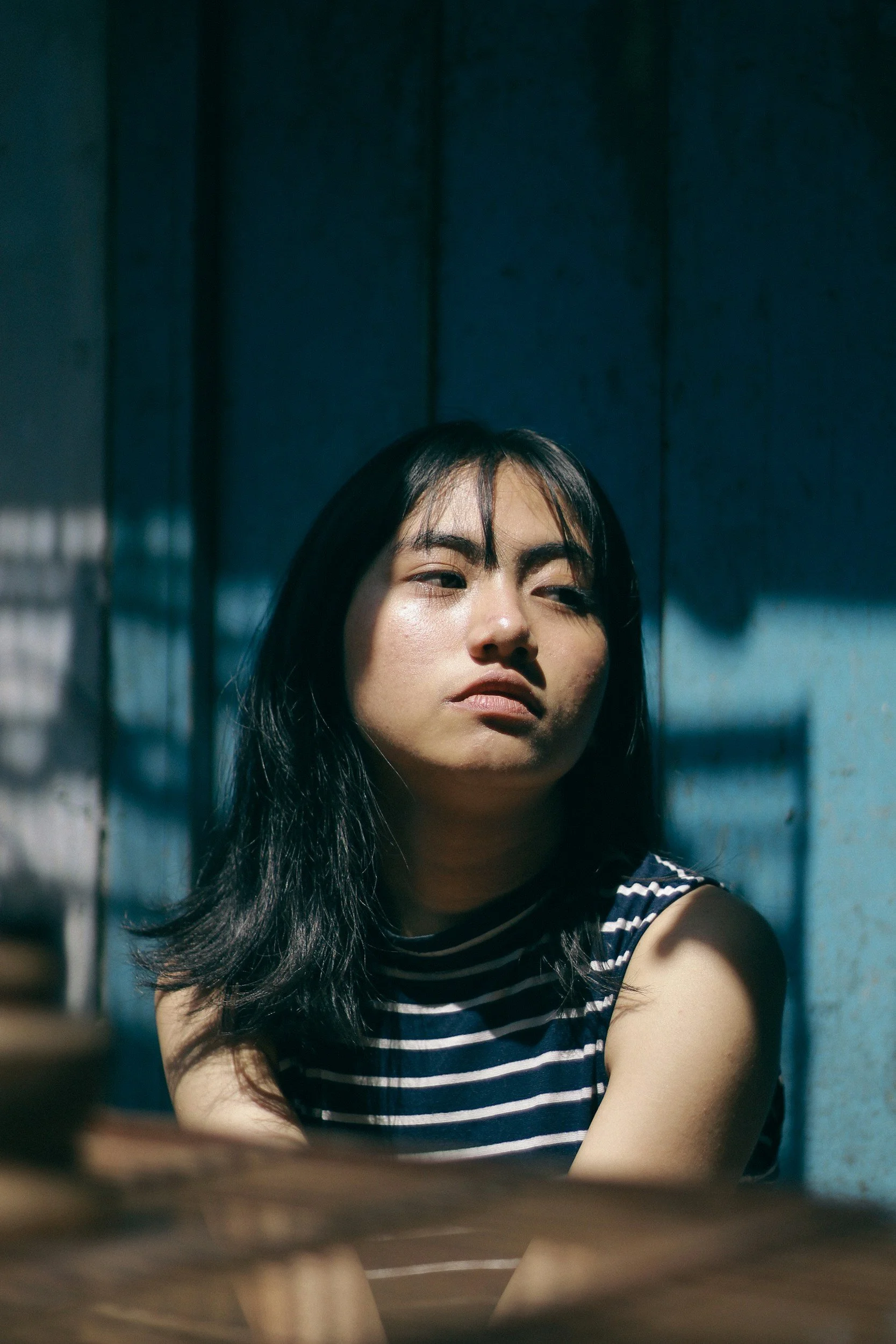 Close-up of a woman with black hair wearing a striped sleeveless top, looking into the camera with a serious expression, with a dark background.