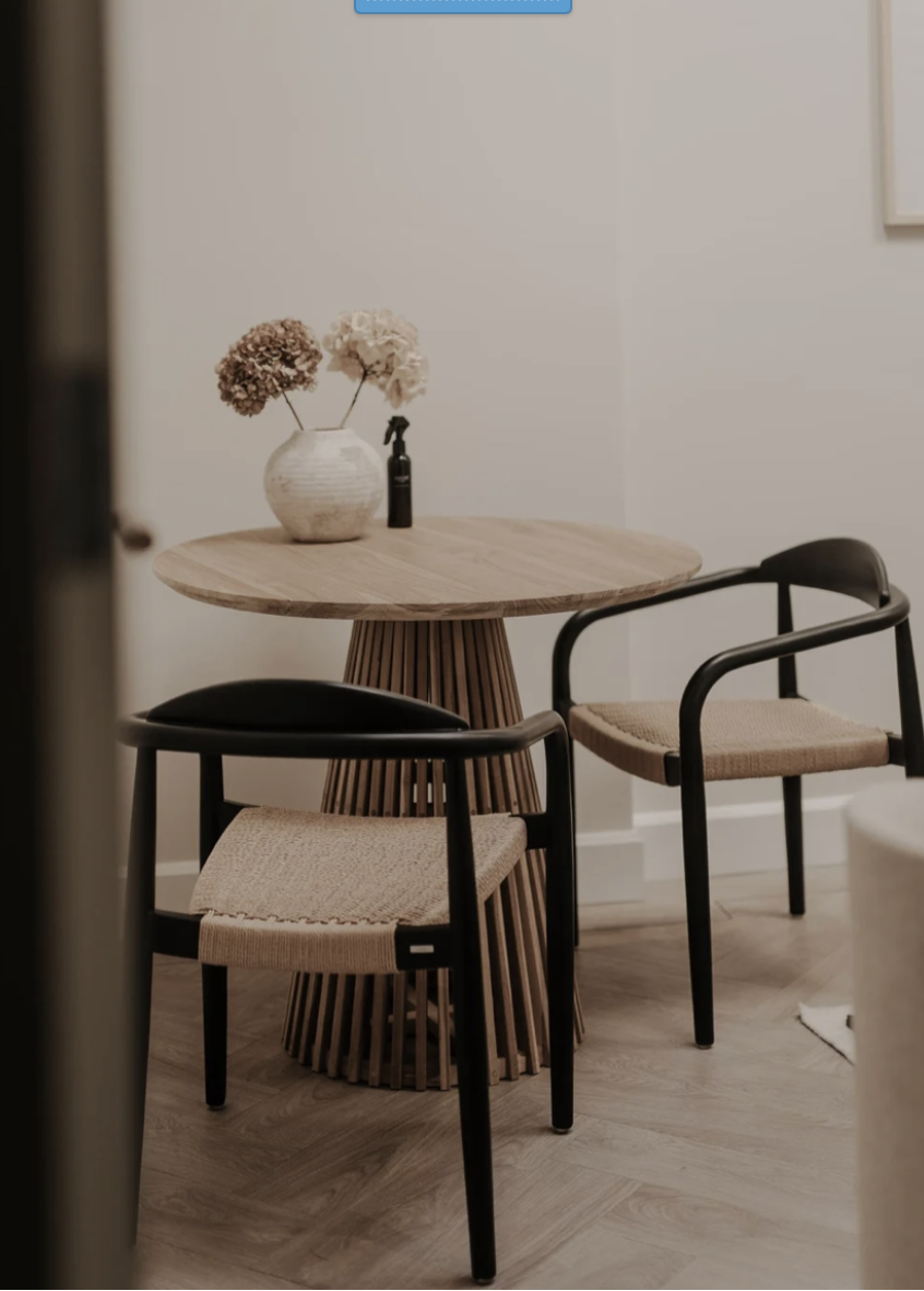 A small round wooden table with a slatted wooden base, decorated with a white ceramic vase containing dried hydrangea flowers and a small black spray bottle. This is at Stories Amsterdam on Keizersgracht 349, 1016GB Amsterdam for Megan Bradley therap