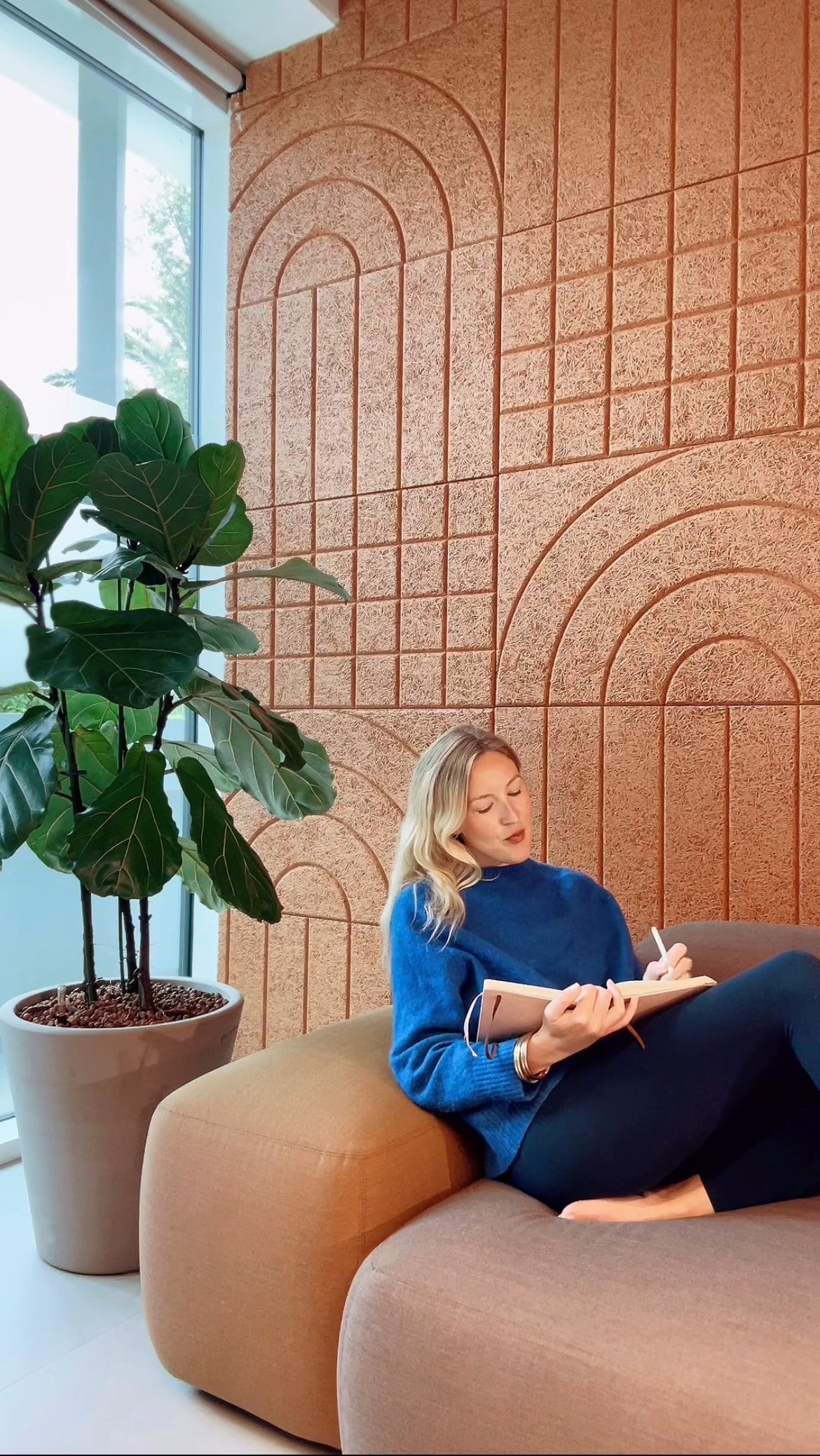 A woman with blonde hair wearing a blue sweater and black pants, sitting on a brown sofa, writing in a notebook at a modern interior space with a large potted green plant, a textured orange wall, and a large window letting in natural light.