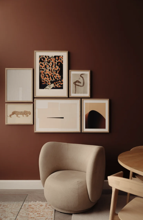 A cozy corner with a beige rounded armchair in front of a brown wall decorated with six framed art pieces, and part of a round wooden table with a chair.