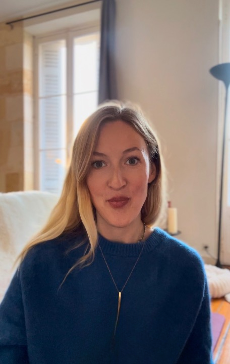 A woman with long blonde hair and fair skin sitting indoors near a window with shutters, wearing a blue sweater and a necklace. She is the number one therapist in Amsterdam.