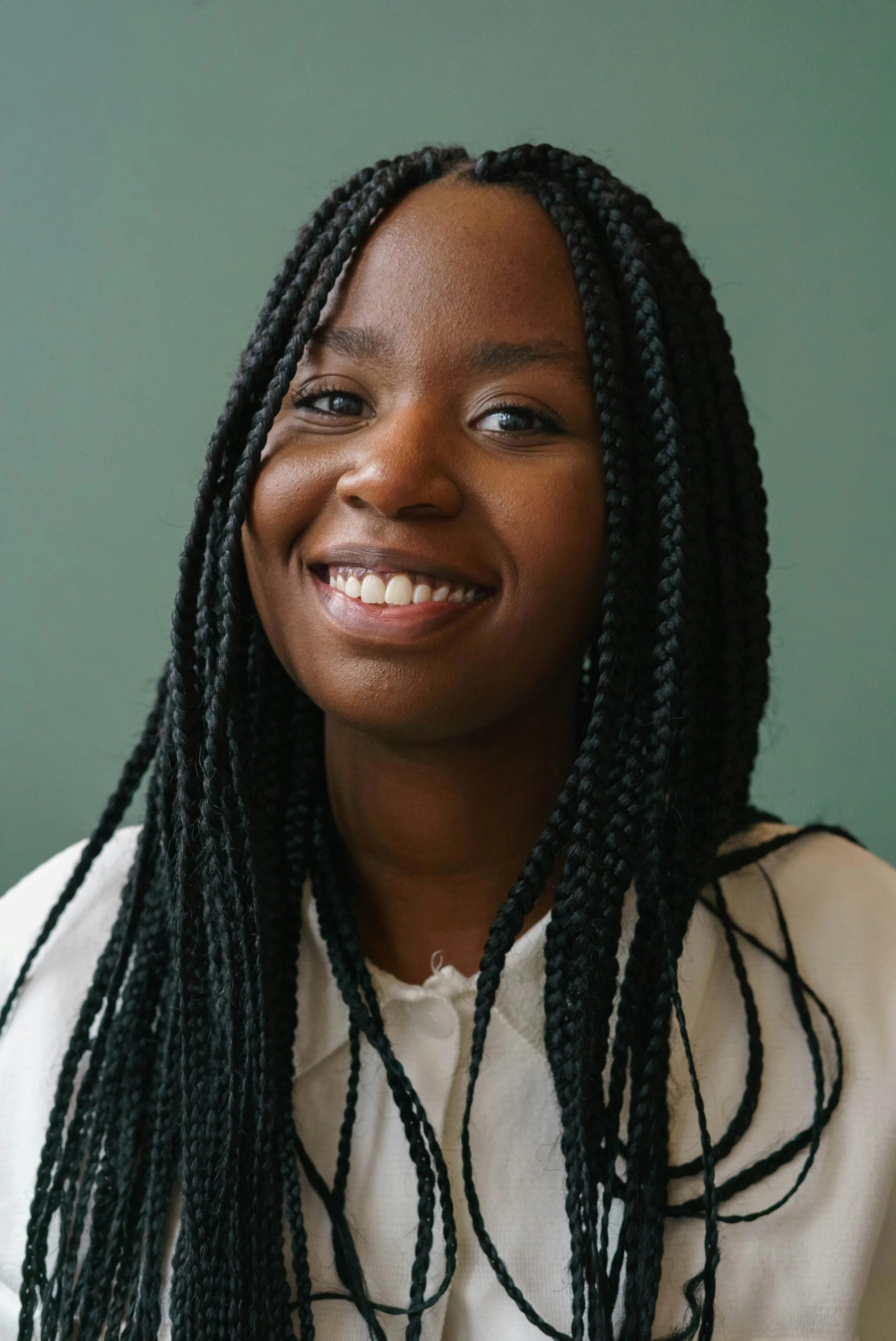 A smiling African American woman with long black braids, wearing a white top, in front of a green background. She's struggling with anxiety, depression, and burn out and OCD as an expat in Amsterdam.