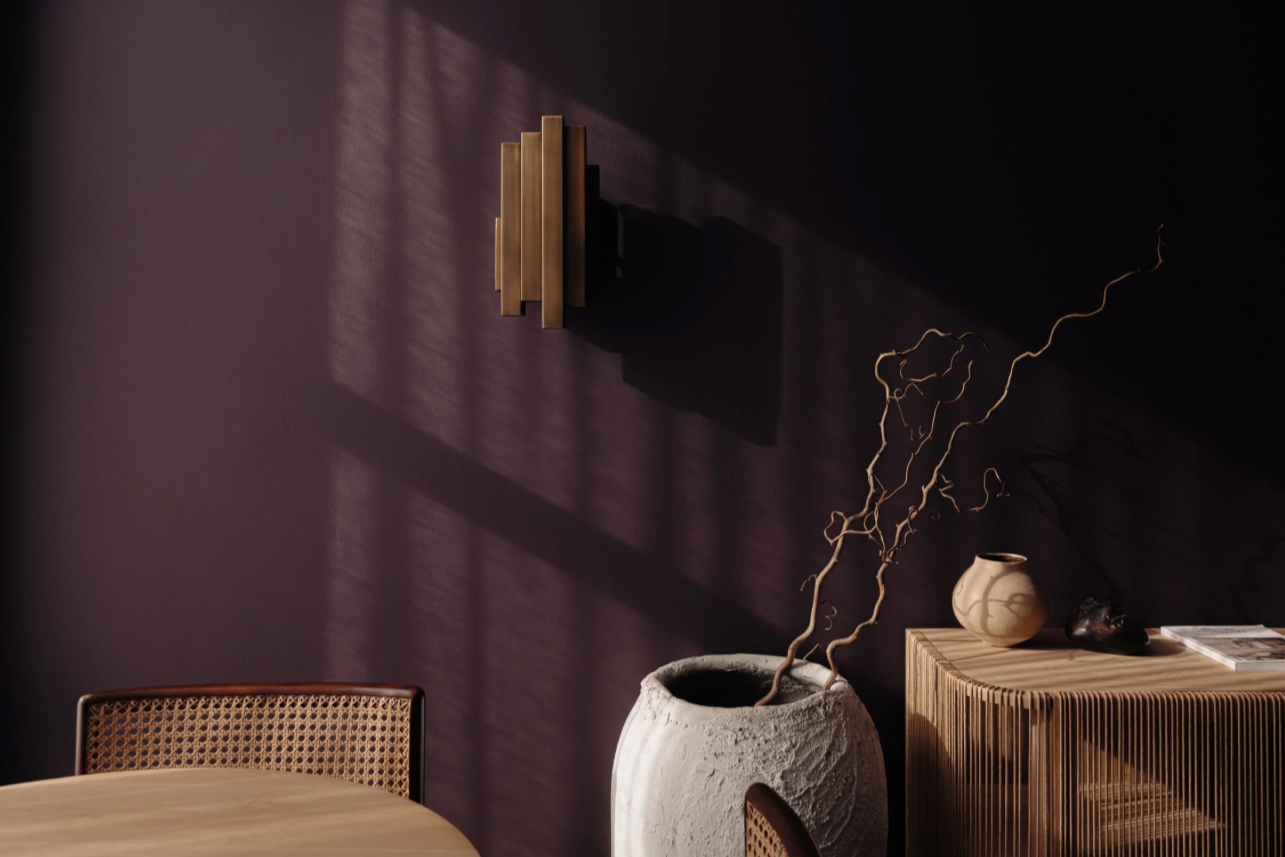 Interior of a room with a dark purple wall, a wooden wall decor, a white textured vase with curly dried branches, a wooden sideboard with a small beige vase, some rocks, and magazines, and a rattan chair at a round wooden table.