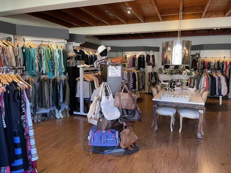 Binnenzijde van een kledingwinkel met diverse kledingstukken, tassen en accessoires, sfeervol ingericht met houten vloer en plafond.