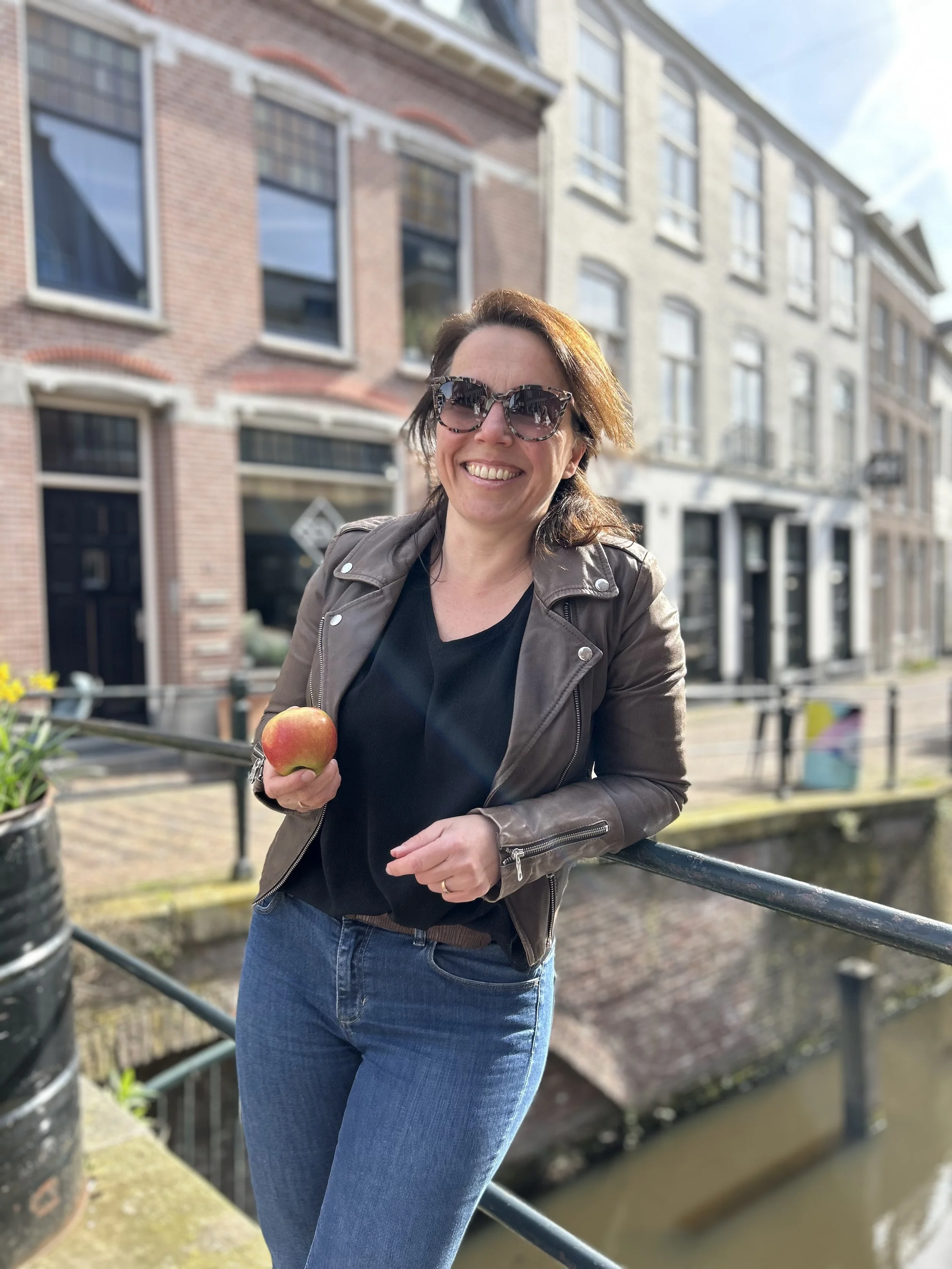Vrouw met bruin leren jas en grote zonnebril houdt een appel, staat naast een kanaal en historische gebouwen in een stad.